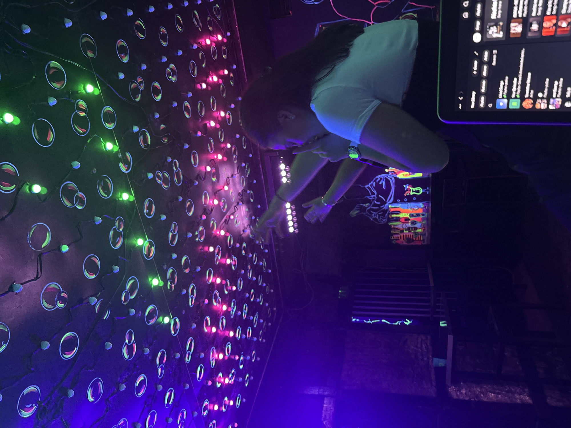 The scene depicts a person in a white shirt interacting with a wall adorned with neon lights and circular shapes. The lights are colored in green, pink, and purple hues. To the right, a tablet displays an interface with multiple app icons and lists. The overall environment is darkly lit, with a visible staircase and brick wall in the background. The location is Unknown, Unknown.