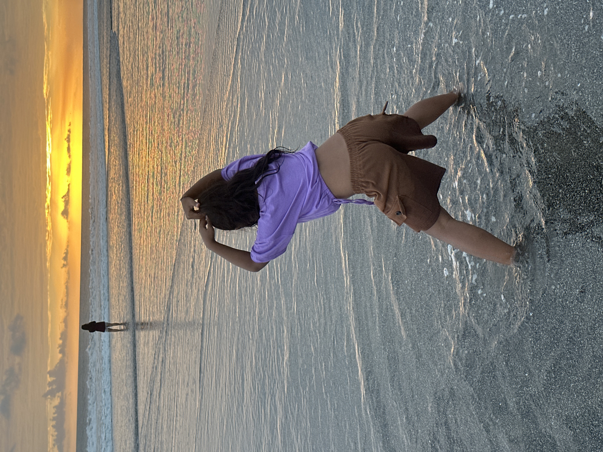 In North Kuta, Indonesia, at approximately 6:30 PM, a person stands in the shallow waters of a beach, facing away from the camera. They are wearing a light purple, cropped t-shirt and brown cargo shorts. Their arms are raised, and their hands are touching their hair. The water is reflecting the warm hues of a sunset, with a bright orange and yellow glow on the horizon and soft, white waves approaching the shore. In the distance, another person stands further out in the ocean, silhouetted against the sunset. The beach itself is composed of dark sand and small pebbles, with shallow water gently lapping at the shore. The overall scene depicts a serene evening at the beach, with the natural beauty of the sunset dominating the backdrop.