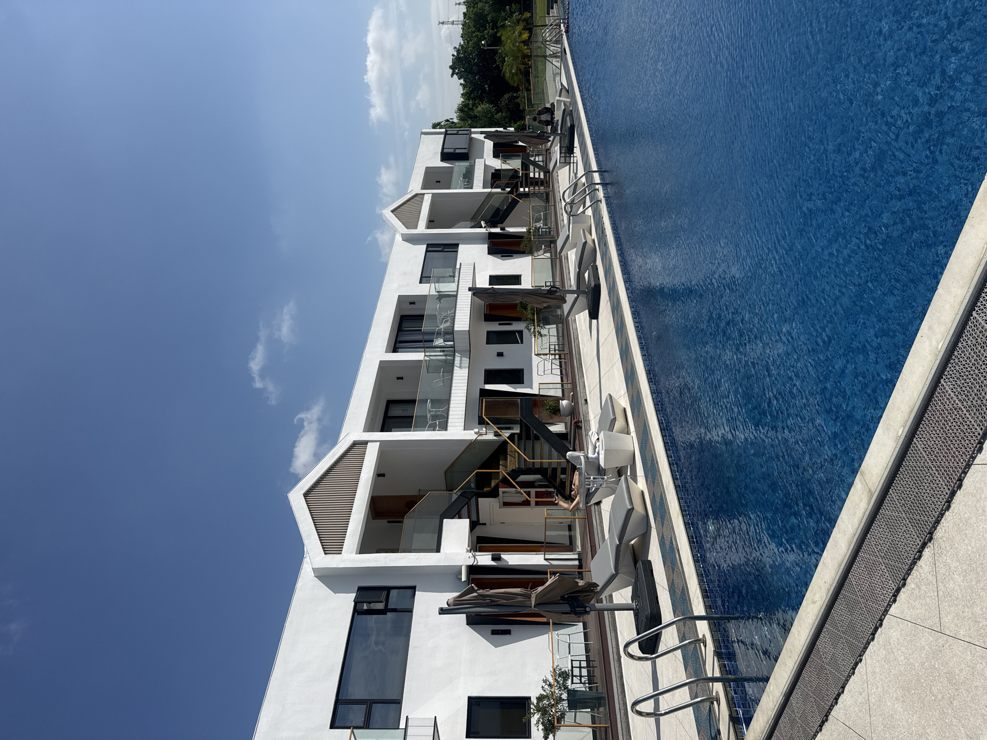 The image depicts a modern, multi-story white building with a geometric design situated beside a large, bright blue swimming pool. The building has multiple balconies with glass railings and dark-framed windows. An outdoor staircase with glass railings and dark steps leads to the upper floors. In front of the building, there are several lounge chairs, umbrellas, and small tables, suggesting a resort or hotel setting. Some of these are arranged on a tiled patio area next to the pool. One person is visible on a lounge chair in the foreground, appearing to relax. The pool has metal ladders for entry. The environment is sunny with a clear blue sky and a few scattered clouds. In the background, there are trees and some distant structures, including what appear to be power lines. The scene is located in Pudong, China.
