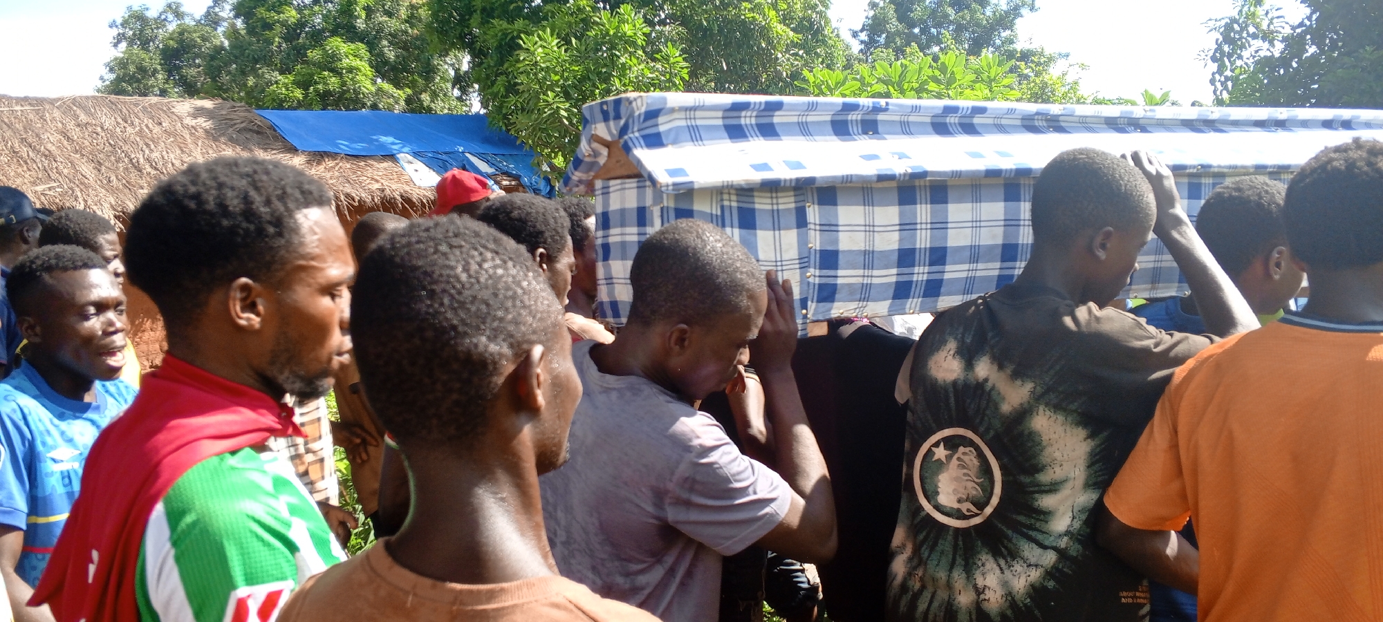 In Ngalula, Democratic Republic of the Congo, a group of approximately ten African men are carrying a coffin draped in a blue and white checkered cloth. The coffin bearers are positioned close together. Some are wearing colored shirts. The environment appears to be a village or rural area, with trees and possibly thatched-roof buildings visible in the background. There is a blue covering over one of the buildings.