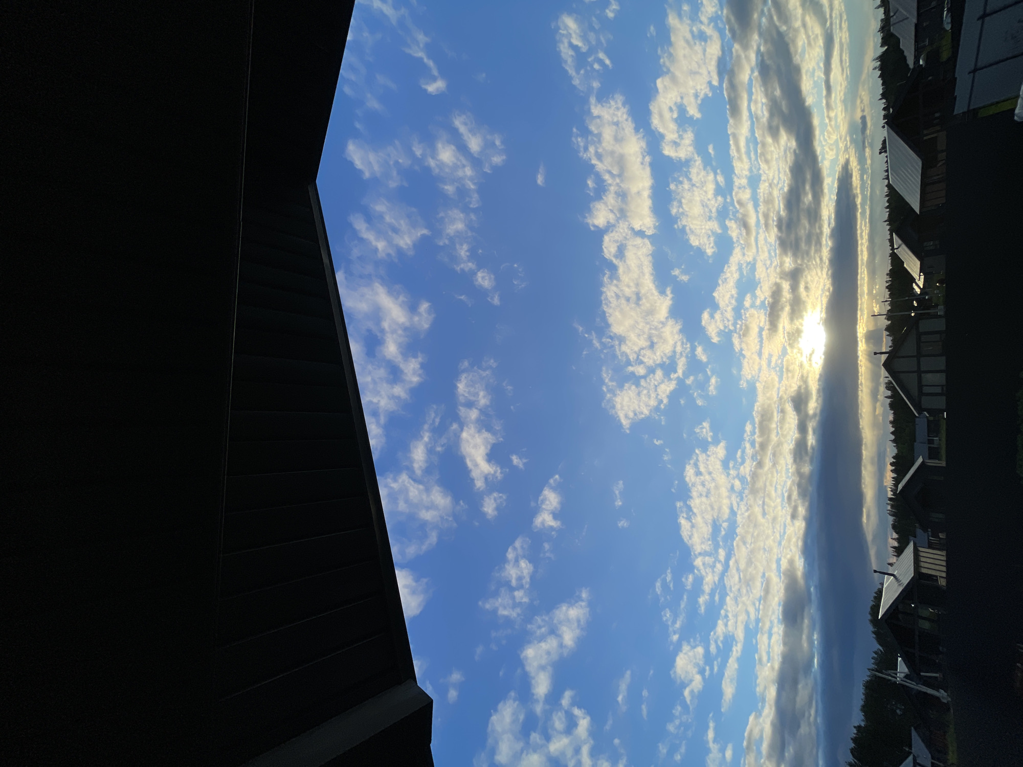 The image captures a view from under a dark, angled overhang, looking out towards a rural or suburban landscape under a bright blue sky with scattered white clouds. The sun is visible near the horizon, partially obscured by clouds, casting a warm glow on the scene. Below the sky, a line of houses with dark roofs and modern architectural styles are visible, interspersed with trees. A dark fence runs across the foreground. The setting appears to be the village of Podshivalovo, Russia, judging by the style of the houses and the surrounding natural environment. There are no people or specific actions depicted in the image.