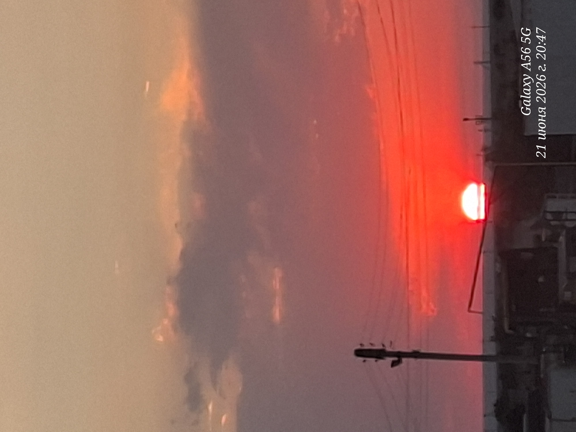 A wide, horizontal, full-shot image captures a vibrant sunset over a rural landscape. The sun, a brilliant orb of white-hot light, is partially obscured by the horizon, casting a fiery red glow across the sky. Wispy clouds, tinged with pink and orange, drift lazily above. In the foreground, silhouetted against the sunset, are several utility poles and power lines, their dark forms stark against the bright sky. In the distance, a low-lying structure and a vehicle are barely visible, suggesting a settlement or industrial area. The overall impression is one of peaceful, natural beauty, with a hint of human presence. There are no people visible in the image.