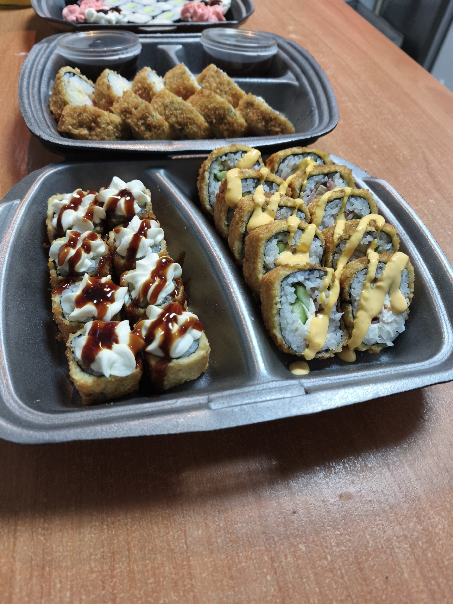 The image displays a close-up, slightly high-angle shot of several trays of sushi, predominantly fried rolls, arranged on a light brown wooden or laminated table.
In the foreground, two black or dark grey partitioned disposable food trays are prominently featured. The tray on the left contains eight fried sushi rolls, each topped with a swirl of white cream cheese and a zigzag drizzle of a dark, glossy sauce, possibly teriyaki or eel sauce. The adjacent tray on the right holds eight other fried sushi rolls, which are generously drizzled with a creamy, yellow-orange sauce, resembling spicy mayonnaise. The cross-sections of these rolls reveal white rice, dark nori, and fillings such as green cucumber and a reddish ingredient, possibly fish or meat.
Further back in the midground, another black or dark grey tray is visible, containing more fried sushi rolls and two small, clear plastic cups filled with a dark dipping sauce, likely soy sauce. In the upper background, out of focus, a fourth tray of sushi is partially visible, featuring rolls with light-colored rice and distinct pink toppings.
The overall scene suggests a takeout or delivery sushi meal, with various types of fried rolls ready for consumption. The lighting is even, and there is no visible text on any of the containers or sushi. The focus is sharp on the foreground sushi trays, gradually softening towards the background.