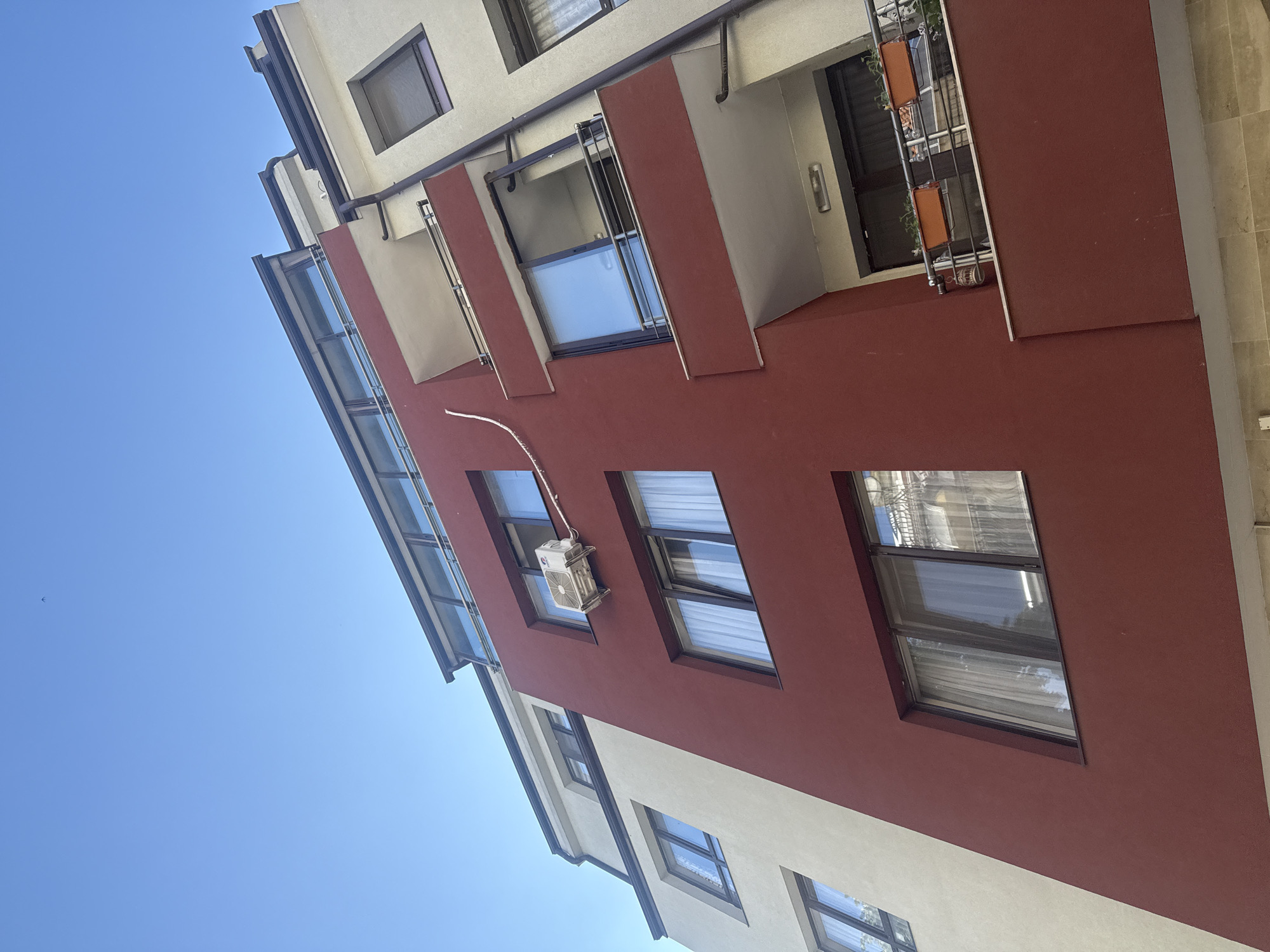 The image is a low-angle shot of a building exterior in Varna, Bulgaria. The building has a modern architectural style with a combination of off-white and red painted sections. Windows are interspersed throughout the facade, with some featuring openable sections. An air conditioning unit is installed near one of the windows, with its connecting cables running along the building's exterior. A balcony is visible on the upper part of the building, with plants or items placed on the railing. The sky is clear and blue, providing a contrasting backdrop to the building's colors.