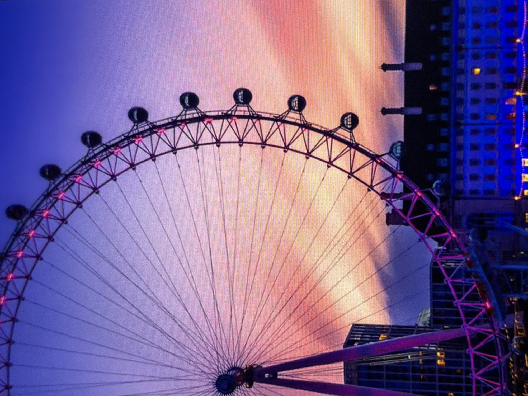 This image captures a large, illuminated Ferris wheel against a vibrant sunset or dawn sky in the city of Maidstone, United Kingdom. The Ferris wheel, featuring a skeletal structure adorned with bright pink and purple lights, dominates the left and upper parts of the frame. Its many spokes radiate from a central hub, extending towards the outer rim where several dark, spherical passenger pods are visible. The sky behind the wheel presents a striking gradient, transitioning from deep blue at the top, through purples, to warm pinks and oranges near the horizon, with subtle cloud formations. In the lower left, the support legs of the Ferris wheel, also bathed in purple light, stand next to a modern building with illuminated windows. To the lower right, a multi-story, traditional-style building with a dark roof and prominent chimneys is visible. Its numerous windows glow with warm interior light, and its lower facade is decorated with cool blue string lights. No individuals are discernible in the scene. The overall impression is one of an urban setting at twilight, emphasized by the interplay of artificial illumination and natural sky colors.