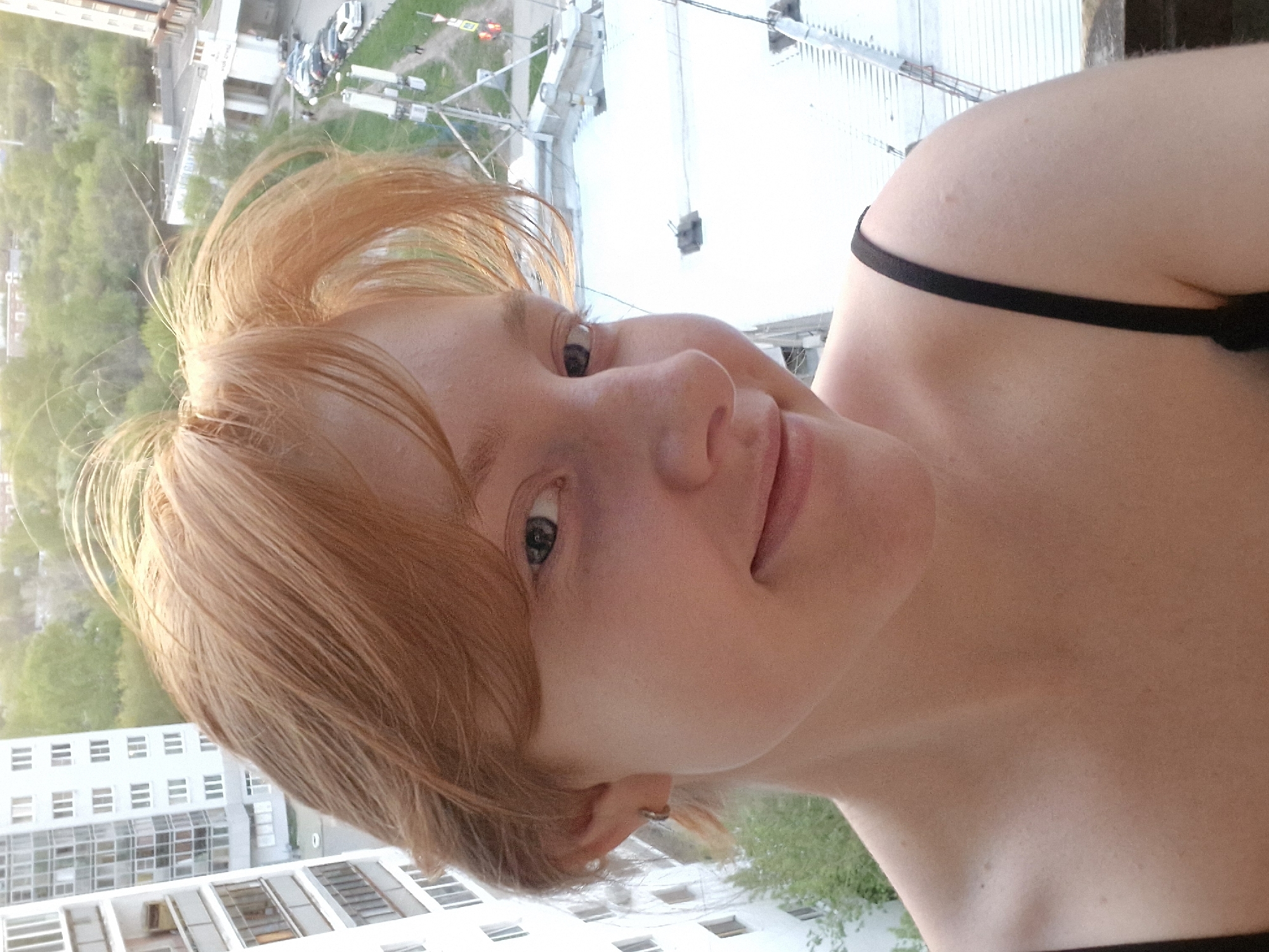 A woman with red hair is positioned in the foreground, likely on a balcony. She is wearing a black tank top. Her head is tilted slightly, and she appears to be looking at the camera. In the background is a cityscape, including apartment buildings, trees, and vehicles. The city is Yekaterinburg, Russia.
