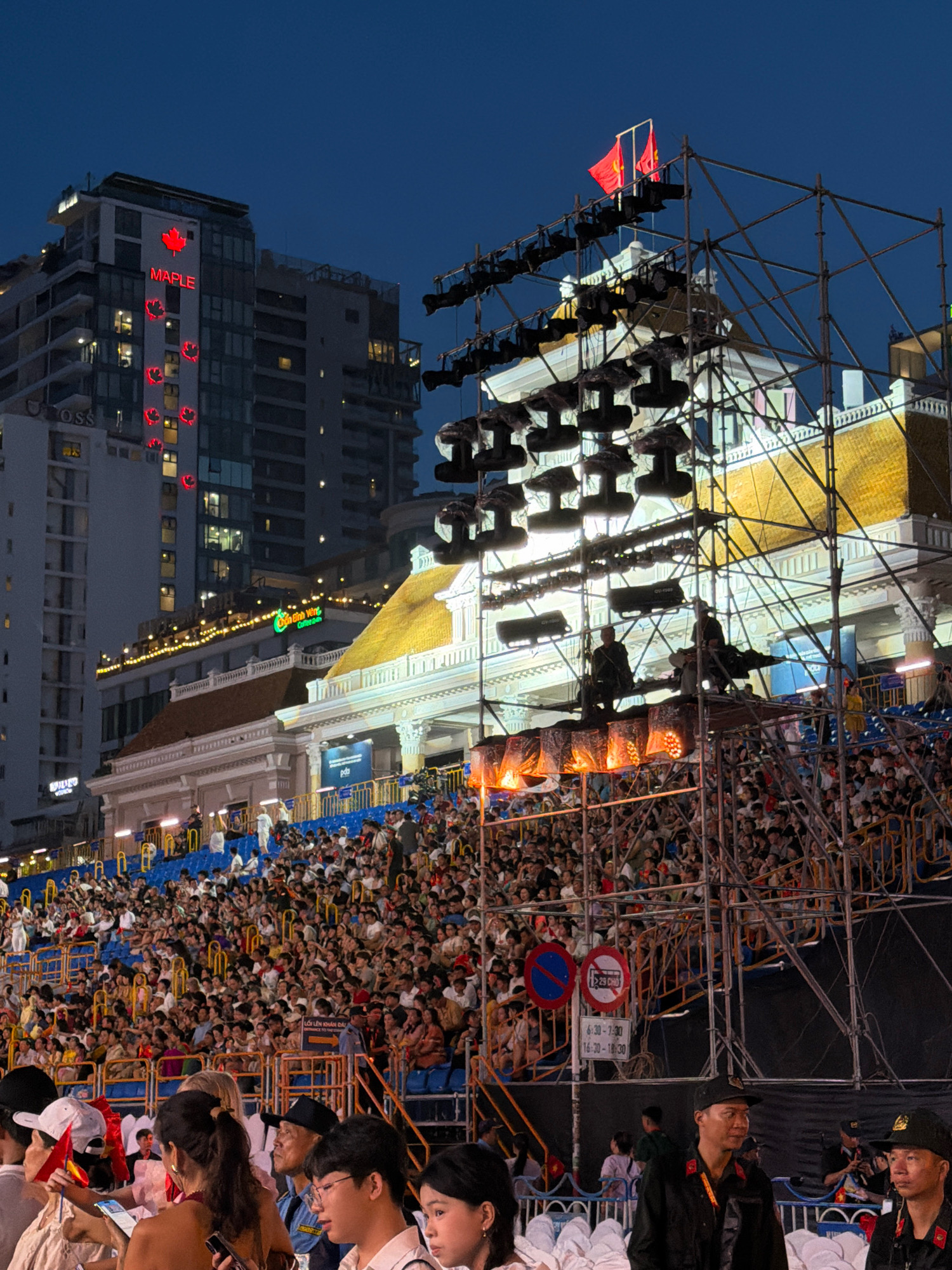 The image depicts an outdoor evening event in Nha Trang, Vietnam, likely a festival or concert, given the large crowd gathered in bleacher-style seating and the extensive scaffolding with lighting equipment. In the background, modern high-rise buildings are visible, one of which features prominent red maple leaf logos and the word "MAPLE" in red neon lights, suggesting a commercial establishment. Two red flags fly atop the scaffolding structure. A large audience occupies the tiered seating area, with many individuals facing the stage area, which is obscured by the scaffolding and lights. The overall atmosphere appears to be festive, with the event taking place after dark, illuminated by artificial lights.