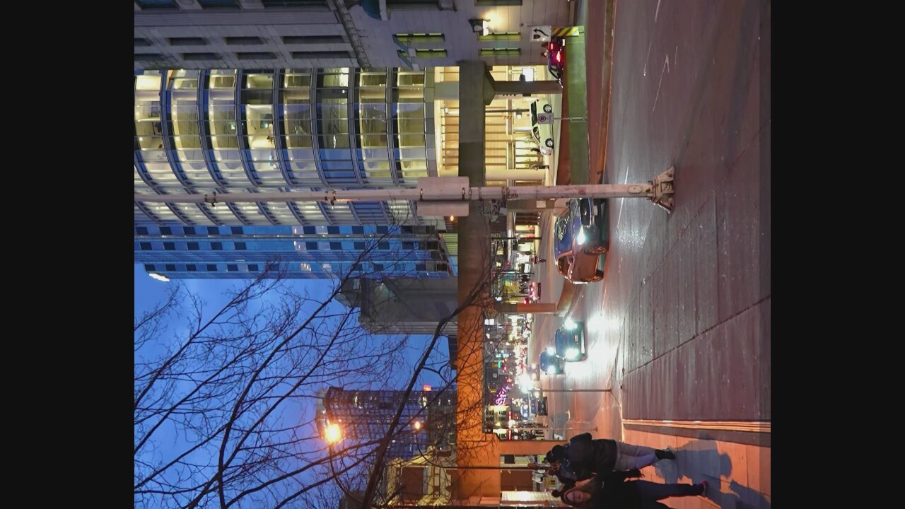 An exterior, medium-angle, eye-level shot shows a city street at dusk. The street is paved and shows a slight sheen from recent rain or dew. Several vehicles are present, moving and stationary. A dark gray sedan is in the foreground, turning left. A dark SUV is behind it, and a lighter colored sedan is further down the street, with its headlights on. The sidewalk on the left has several pedestrians walking. Tall buildings with many windows line the street, some illuminated from within, indicating offices or residences. A bare-branched tree is visible in the upper left quadrant. Overhead, a concrete pedestrian walkway spans the street. The sky is a deep twilight blue. A lamppost stands on the right side of the street.