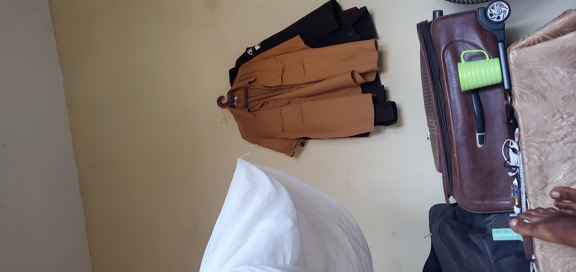 The image depicts an indoor scene in Numan, Nigeria, with a wall as the backdrop. A tan-colored short-sleeved shirt with two front pockets is hanging on a purple hanger against the wall. Behind it, a black garment, possibly a jacket, is also hanging. To the left of the shirt, a large piece of white fabric, likely a mosquito net or sheet, is draped. In the foreground, a brown suitcase with a black handle is placed on a table covered with a textured, patterned cloth. On top of the suitcase, a bright green ribbed mug is positioned next to a black object, possibly a phone. A person's foot, with visible toes and dark skin, is visible in the lower left corner of the image, resting on the patterned cloth. Various small items, including a pen, earbuds, and a rectangular object, are scattered on the suitcase.