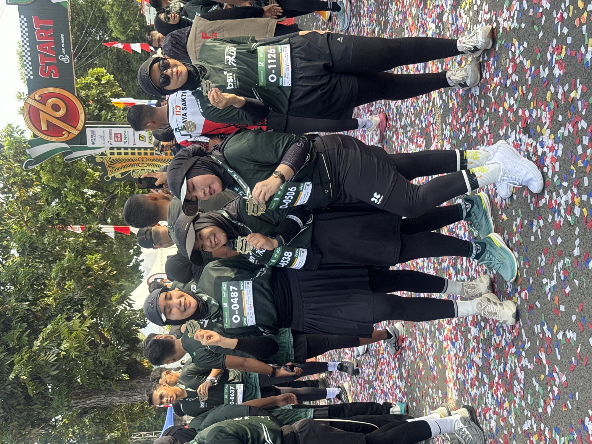 The image depicts a group of people, predominantly women wearing matching dark green t-shirts and headscarves, standing on a street covered in confetti. The event appears to be a running race, possibly a marathon, with participants displaying medals around their necks and bib numbers on their shirts. The bib numbers are for a 5K race, with visible numbers like "O-0487", "O-0506", and "O-1126". In the background, a banner with "76 START" and "JAK PLUENG" is visible, suggesting the start or finish line of the race. The environment is outdoors, with trees and tents in the background, indicating a public event. The people are posing for a photograph, smiling and holding up their medals. The presence of the "76 START" banner and the mention of "JAK PLUENG" in the context of a running event suggests the location is likely related to a significant anniversary or celebration, possibly within Lhokseumawe, Indonesia, given the context of previous analyses.