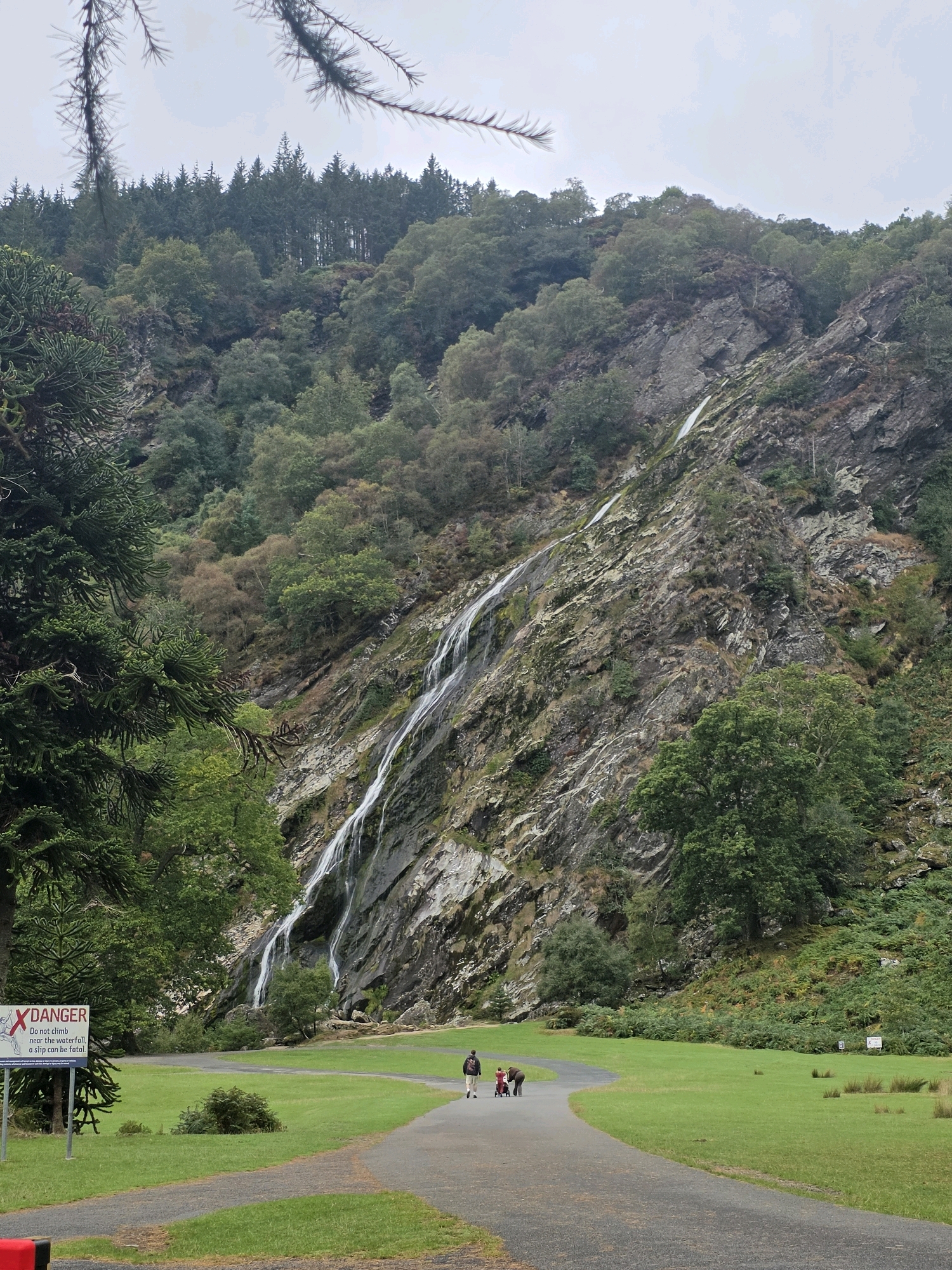 The image shows a tall, cascading waterfall flowing down a rocky mountainside in Old Long Hill, Ireland. The waterfall is surrounded by lush green trees and vegetation. In the foreground, a paved path winds through a grassy park. A sign with a red "X" and the word "DANGER" is visible on the left side of the path, warning people not to climb near the waterfall. Three people, one adult and two children, are walking away from the camera on the path. The sky is overcast, and the overall mood of the scene is serene and natural.