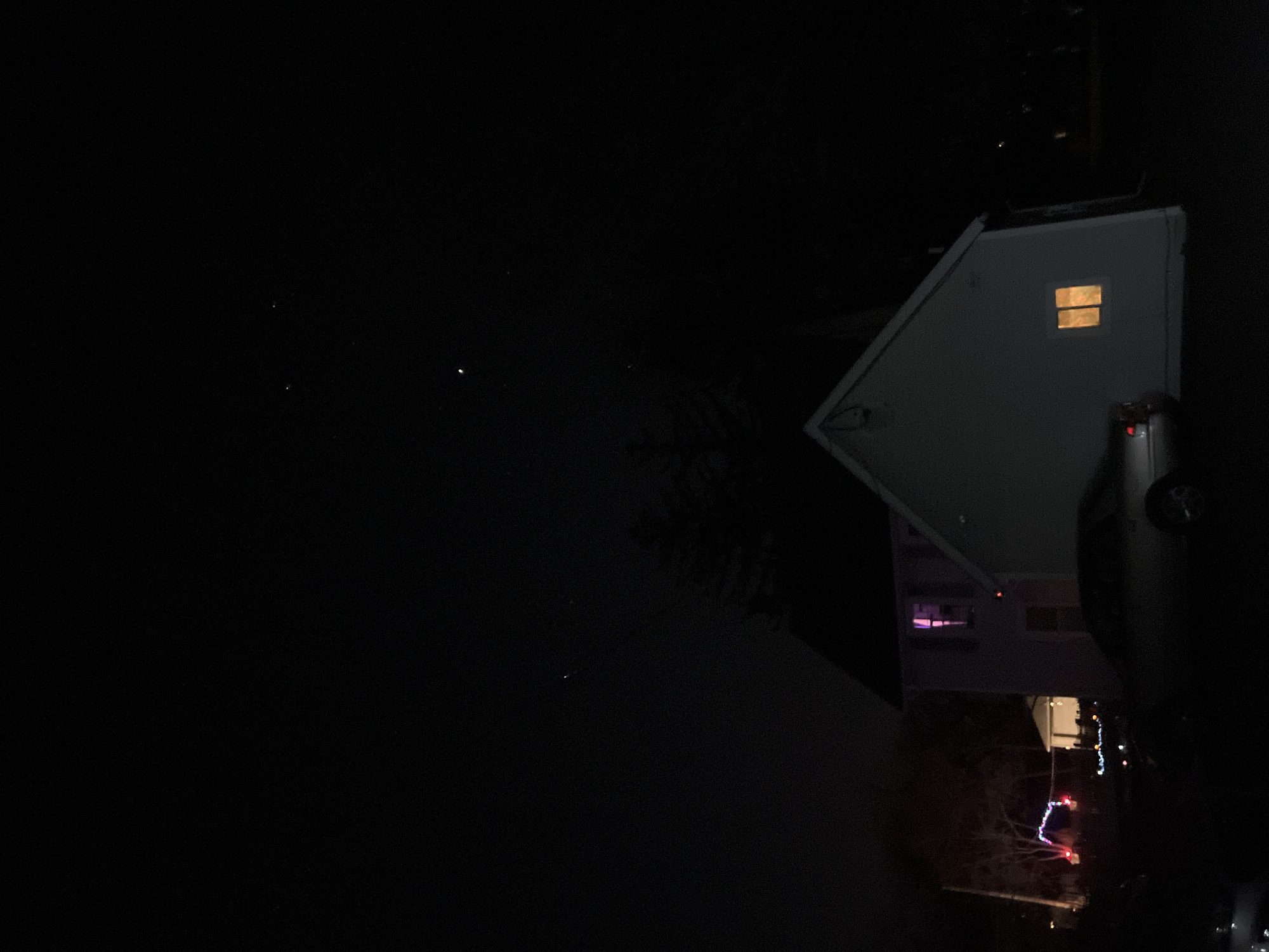 The image, taken at night in the City of Newburgh, United States of America, shows a house. The house is gray with an illuminated window. Adjacent to the house is a vehicle. The image shows Christmas lights in the yard.