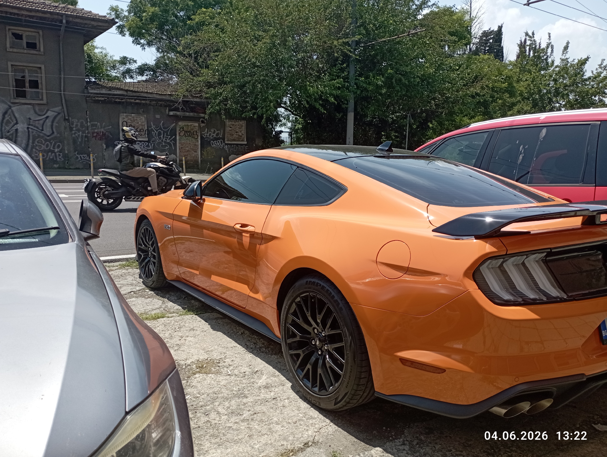 The image captures an outdoor urban or semi-urban scene on a bright, sunny day. The primary subject is an orange Ford Mustang coupe, parked with its rear and passenger side prominently displayed. The Mustang features black multi-spoke wheels, a black roof, and a black rear spoiler. Text "5.0" is visible on its side fender, and the tire sidewalls show "IMPERIAL", "ECOSPORT", and the size "275/40R19". A partial license plate starting with "BG" is visible on the rear.
In the immediate foreground, the front of a silver car is partially visible, obscuring part of the Mustang. To the right of the Mustang, a red SUV or minivan is partially visible in the background.
Further in the background, a motorcyclist wearing a helmet, dark jacket, and light-colored pants is riding a black motorcycle. Behind the motorcyclist are old, dilapidated buildings with broken windows and extensive graffiti, including discernible words like "SOLO BOMB" and "Hobo!". Overhead, lush green trees and power lines are visible against a clear blue sky.
The scene is well-lit by natural sunlight, indicating midday or early afternoon. A digital timestamp "04.06.2026 13:22" is visible in the bottom right corner of the image.