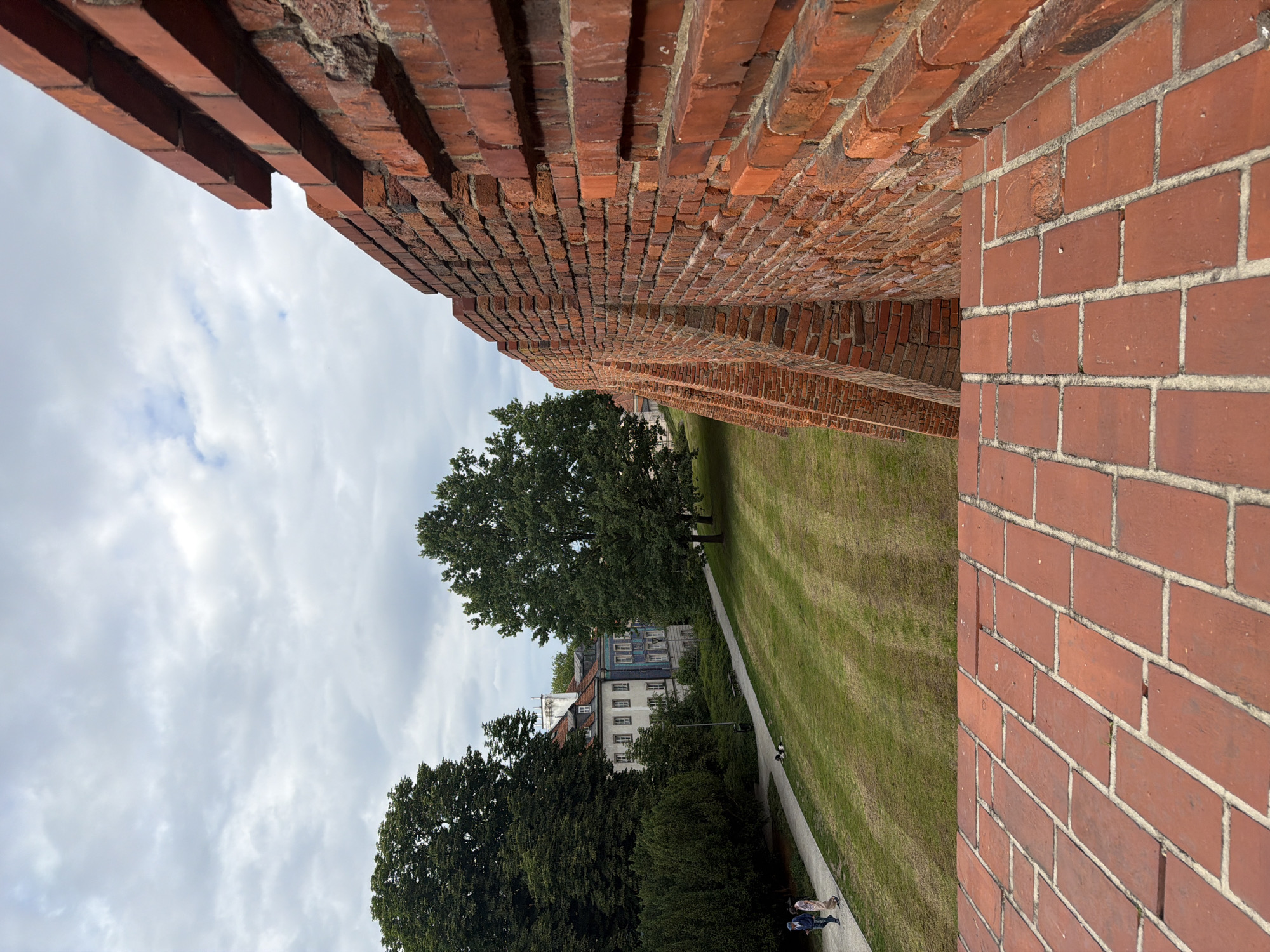 A close-up, right-angled perspective from a high vantage point shows a weathered brick wall in the foreground. The wall is made of red bricks, with some showing signs of age and wear. Its surface is rough and textured, and it appears to be part of a historical structure. Beyond the wall, a grassy park slopes downwards, with a paved walkway running along its edge. Two people, a man and a woman, are walking on the path. In the background, several buildings with pitched roofs are visible, nestled amongst lush green trees. The sky above is overcast with grey clouds.