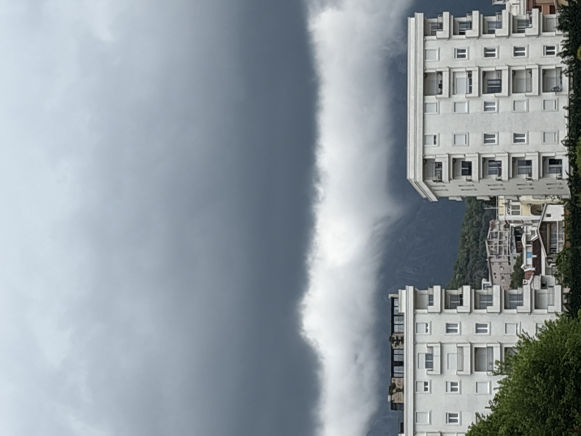 This dramatic landscape photograph captures two light-colored buildings set against a striking atmospheric condition, likely in Bar, Montenegro. The image is dominated by a sharply defined horizontal band of bright, dense cloud or fog that stretches across the middle ground. To the left and above this band, the sky is a uniform, dark, heavy gray, suggesting a deeply overcast sky or an approaching weather front. To the right of the cloud band, and partially obscured by it, a mountainous or hilly background can be seen.
In the lower right portion of the frame are two prominent, multi-story buildings, likely apartment blocks or hotels. They feature a minimalist, modernist architectural style with numerous windows and balconies, appearing light-colored against the darker background. Lush green trees and foliage are visible in the immediate foreground, particularly on the right side of the lower building. Hints of other structures are visible on the hillside behind the main buildings.
No people are visible, and no specific activity is taking place; the scene is a still landscape. The time of day appears to be daytime, but the heavy cloud cover gives the scene a somber or atmospheric quality. There is no visible text in the image.
