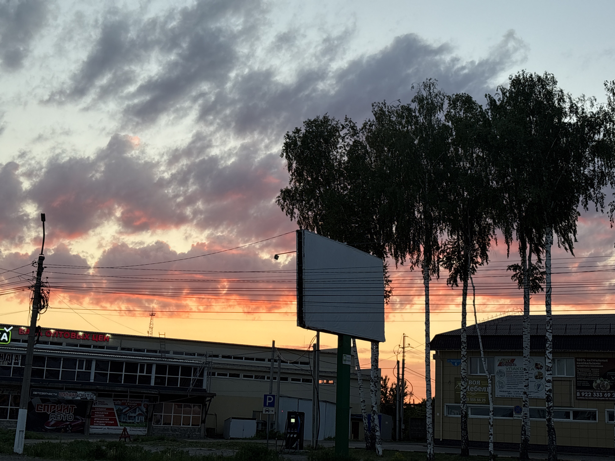 The image depicts a street scene in Chaikovsky, Russia, at sunset. A large blank billboard stands near the center of the frame, silhouetted against the colorful sky. In the background, there is a long, low building with a sign that reads "БА - ОПТОВЫХ ЦЕН," as well as signs for "Автозапчасти СПРИНТ АВТО".
A smaller building to the right has birch trees in front of it and posters advertising "УПАКОВКА" and "100% НАТУРАЛЬНЫЕ" products. Utility poles and wires are visible overhead, running across the sky. The sky displays a gradient of colors, with orange and yellow hues near the horizon fading into purple and gray clouds above. A parking meter and signage are visible near the front of the first building.