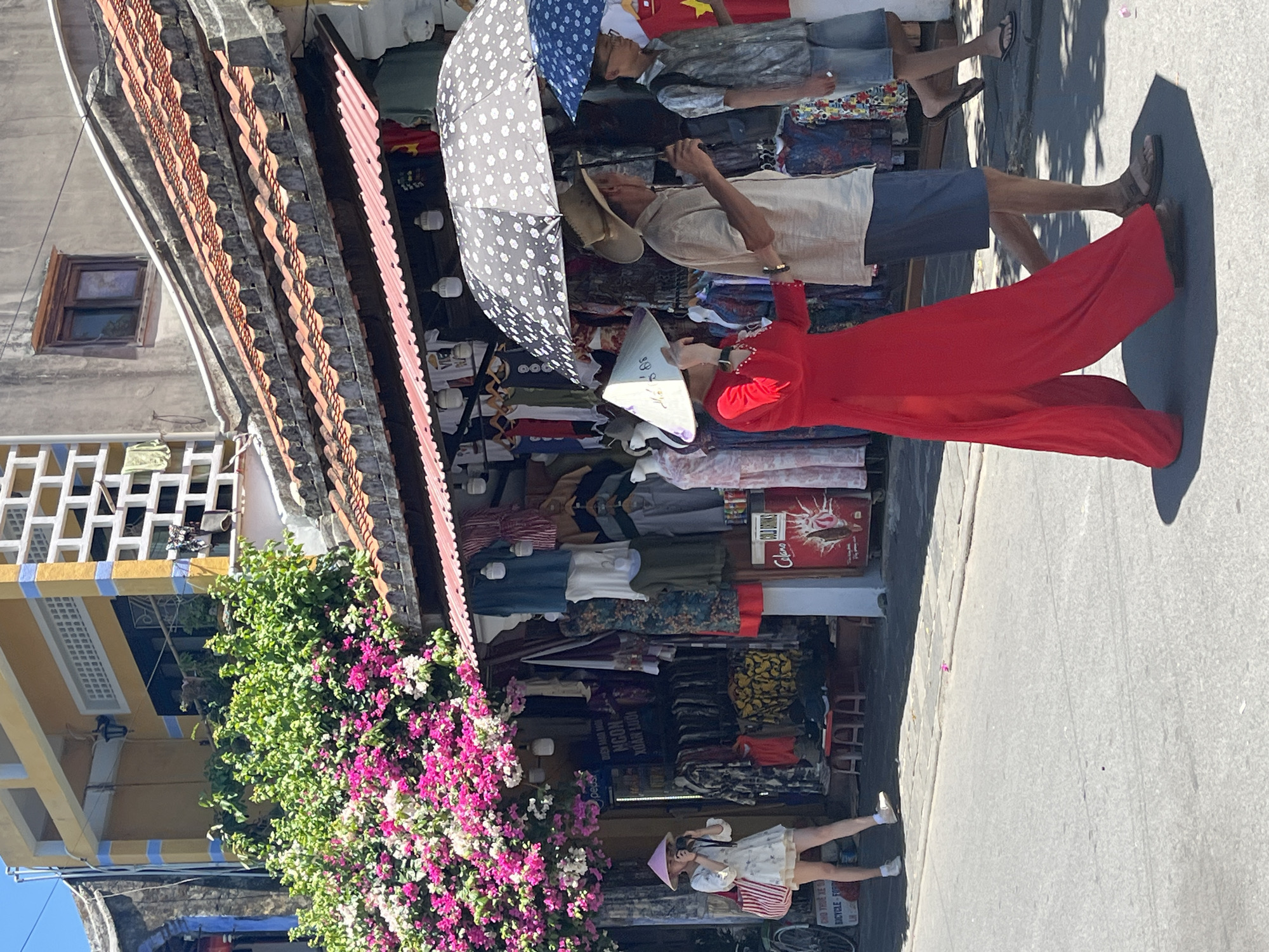 This image captures a vibrant, sunlit street scene in Hội An, Vietnam, bustling with people and market activity. The view looks along a paved street lined with market stalls on the left side.
In the mid-ground, a woman wearing a striking red áo dài (traditional Vietnamese long dress) and a nón lá (conical hat) stands on the paved path, casting a long shadow that indicates bright sunshine. She appears to be looking towards the market stalls or down the street. To her left, market stalls are shaded by awnings and large umbrellas, including a prominent grey one with white polka dots and a blue one. Behind these, a man in a straw hat is visible, seemingly using a phone, and another man in a t-shirt and jeans is under one of the umbrellas. The stalls are filled with various garments displayed on racks and shelves.
On the far left, a multi-story building with a yellow facade and blue striped trim is partially covered by lush green foliage adorned with vibrant pink and white bougainvillea flowers. Below the main market area, towards the bottom left, a younger woman in a white dress and a pink nón lá is seen holding a camera, possibly taking a photograph, with a bicycle parked nearby. Architectural details like terracotta roof tiles are visible above the stalls.
The time of day appears to be mid-day due to the strong shadows. Visible text includes "COLD DRINKS" and "Coca-Cola" on a red sign, "BICYCLE FOR RENT" on a white sign, and "PEPSI" on another small sign. Vietnamese text "HỘI AN MOON" and "XUÂN LƯU" are partially visible on a dark sign behind the market stalls. The scene depicts a lively market environment, typical of a sunny day in Hội An.