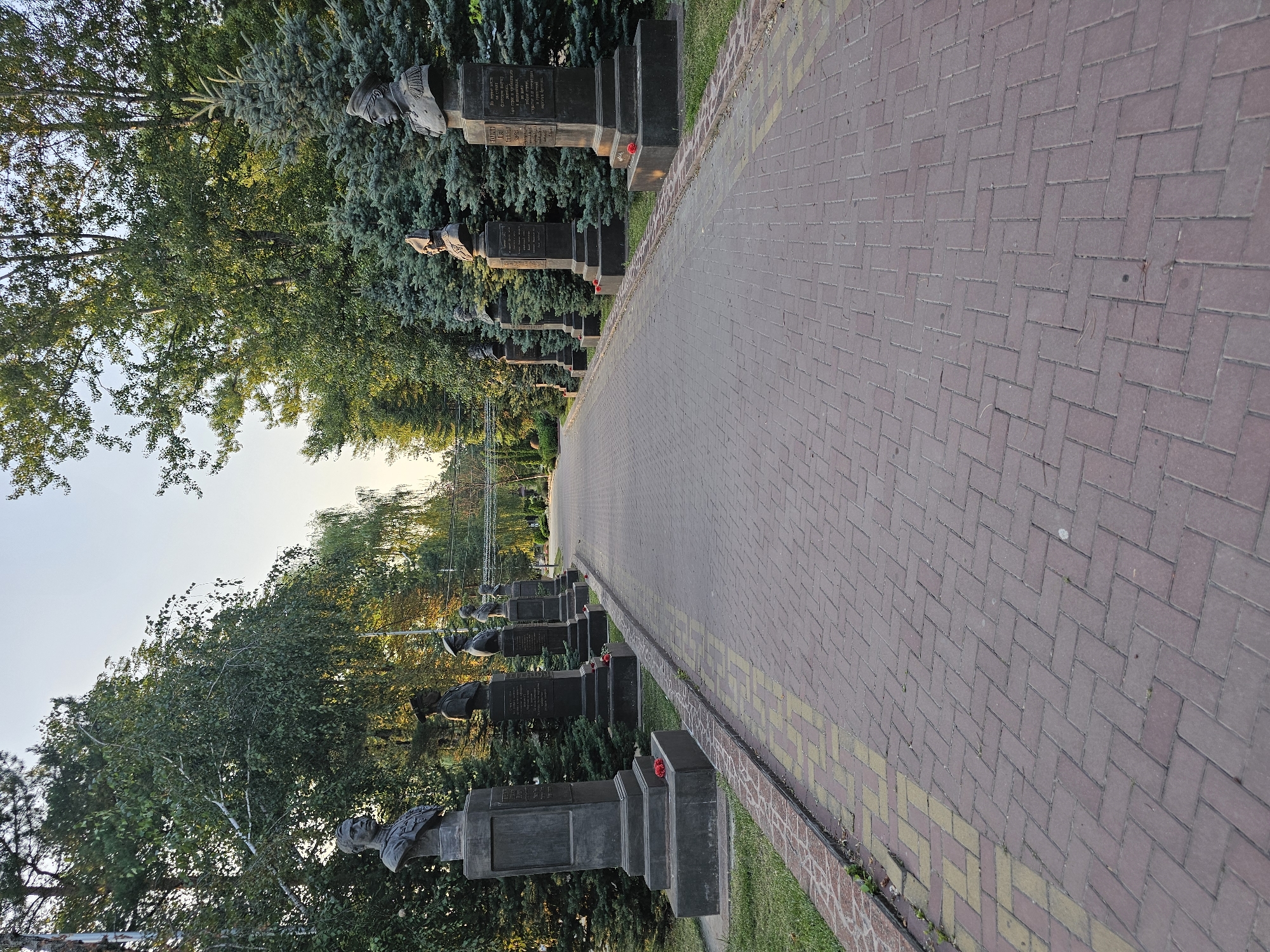 The image shows a pathway in a park lined with busts of historical figures. The pathway is paved with reddish-brown bricks laid in a herringbone pattern, with a yellow border. On either side of the pathway, there are manicured green lawns, tall trees, and dark green fir trees. Bronze busts of men in military uniforms are mounted on tall, dark grey stone pedestals. Each pedestal has an inscription. Red flowers are placed at the base of several of the pedestals. The sky in the background is a pale blue, suggesting it is either early morning or late afternoon. The overall scene is one of a solemn memorial or historical landmark.
This scene depicts the Alley of Heroes in the city of Cherkessk, Russia.