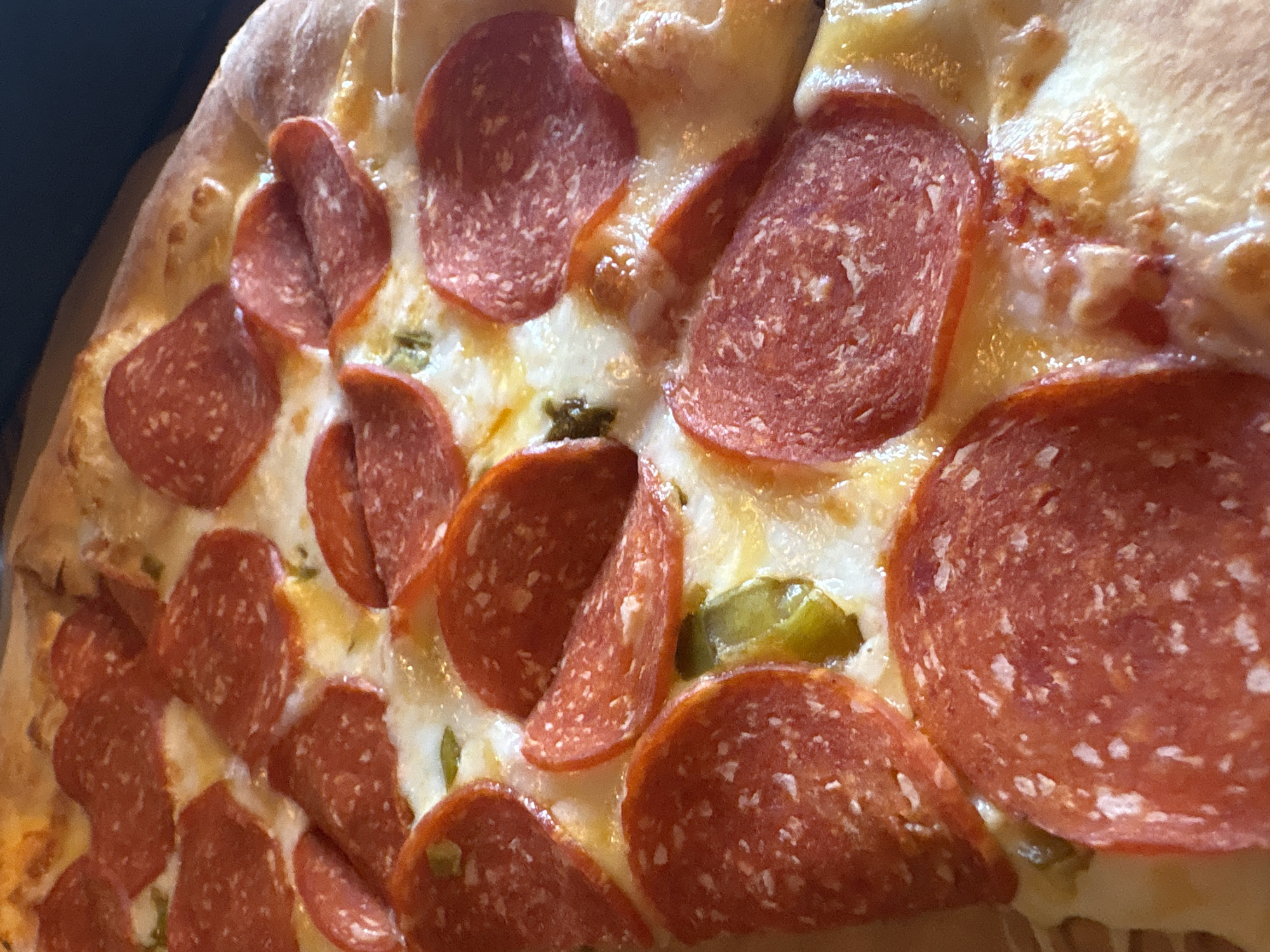 A close-up, high-angle shot captures a delicious-looking pepperoni pizza. The focus is tightly on the toppings and a portion of the crust, filling the majority of the frame. Numerous slices of bright red pepperoni are generously scattered across a bed of melted, bubbly, golden-white cheese. The pepperoni slices exhibit slightly curled edges, a common characteristic after baking. Interspersed among the cheese and pepperoni are small, chopped pieces of green vegetable, likely bell peppers or jalapeños, adding a touch of contrasting color.
The pizza crust is visible along the left and top edges of the frame, appearing golden brown and slightly puffed, suggesting a well-baked dough. The texture of the cheese is visibly stringy and glossy in areas, indicating its freshly melted state. The lighting is soft and warm, highlighting the textures and colors of the pizza ingredients. The background is dark and out of focus, suggesting the pizza is resting on a surface or in a box, drawing all attention to the food itself. There are no people visible, and no specific activity beyond the presentation of the pizza. No visible text is present in the image. The scene is likely indoors, possibly in Perm, Russia, as per the provided context.