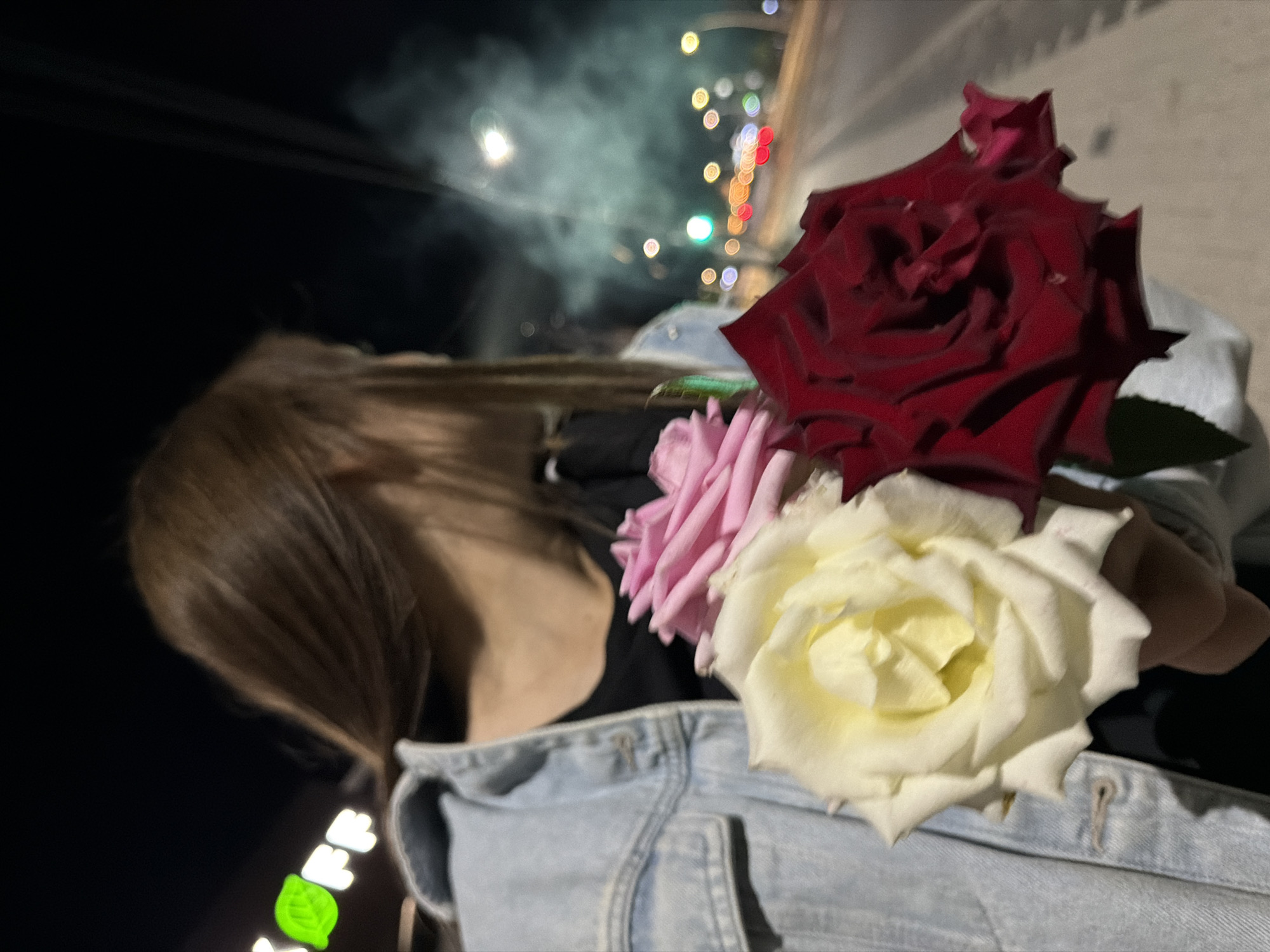 The image shows a person from the shoulders down, holding three roses: one white, one pink, and one dark red. The person is wearing a light blue denim jacket over a dark shirt. The background is dark, suggesting it is nighttime. Out-of-focus lights from vehicles and streetlights are visible, indicating the scene is on a street or roadside. A green and white sign, possibly a business logo, is partially visible to the upper left of the frame. There is also a faint haze or smoke-like phenomenon in the upper right portion of the background. The context of Strășeni, Moldova, is external to the visual information provided in the image.