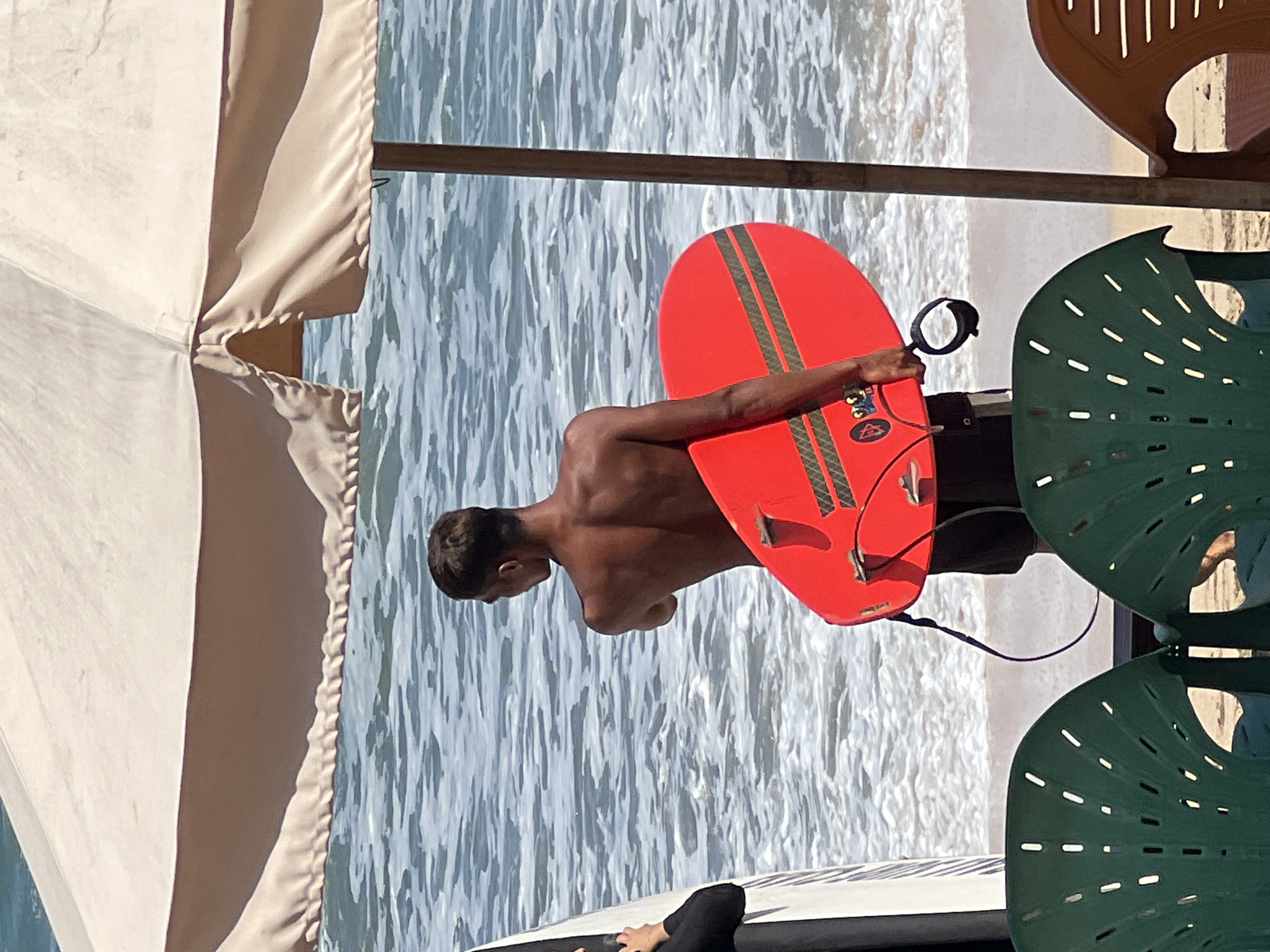 In Kuta, Indonesia, a young man stands on a beach with his back to the camera, holding a red bodyboard. He is shirtless, wearing dark shorts, and his muscular back is visible. The bodyboard has two dark green stripes and a black grip leash attached. To his left, another person wearing a black wetsuit is partially visible, also holding a white surfboard. In the foreground, two green plastic chairs are positioned on the sand, facing the ocean. A large, light-colored beach umbrella shades the scene. The ocean is visible in the background, with gentle waves breaking on the shore. The sun is shining, casting shadows and highlighting the textures of the sand and water.