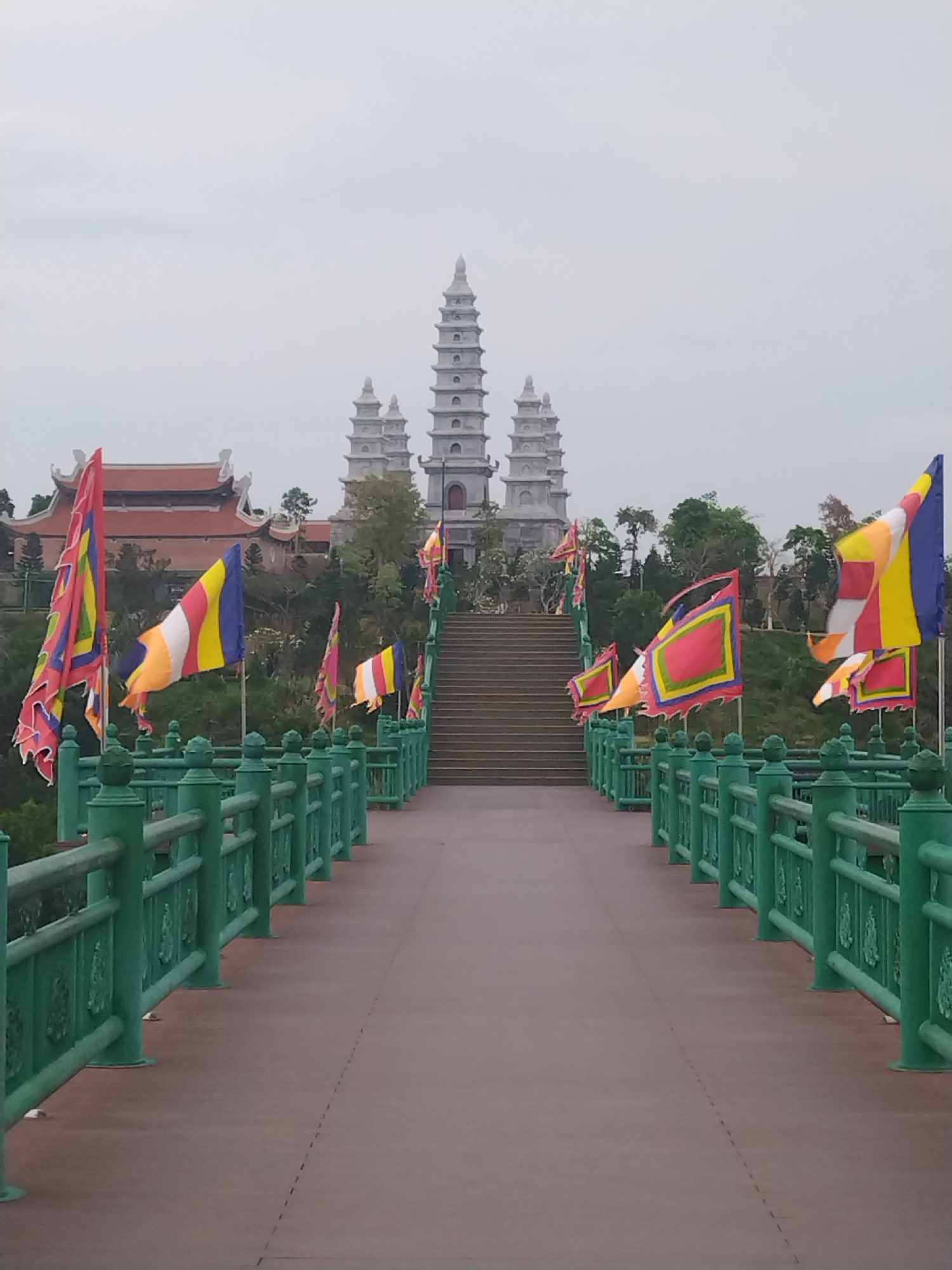 The image depicts a bridge leading to a complex of religious buildings in Hong Gai Ward, Vietnam. The bridge has a reddish-brown surface with green railings adorned with decorative elements. Several brightly colored flags are attached to the railings on both sides of the bridge. In the background, a building with a red tile roof is visible to the left, and a cluster of tall, tiered white pagodas rises in the center of the frame. Green trees and foliage surround the buildings. A series of steps leads up to the pagodas. The sky is overcast.