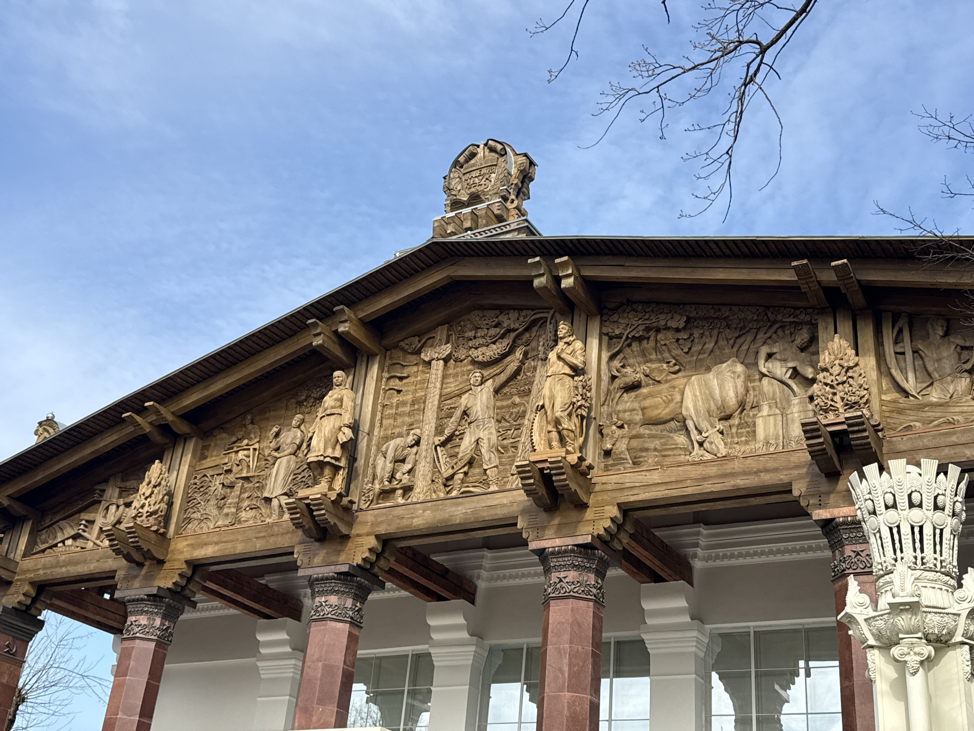 The image depicts the facade of a building in Moscow, Russia. The architecture is characterized by ornate wooden carvings depicting scenes of agricultural life and socialist themes. The building's pediment is adorned with figures of workers, farmers, and animals such as cattle, all intricately carved into the wood. The roof is topped with a decorative element bearing what appears to be Russian text. The supporting columns are a combination of maroon-colored, possibly stone or concrete, and ornate white columns with decorative capitals. The sky is a light blue with scattered clouds, and bare tree branches are visible in the upper right corner. The building appears to be well-maintained and is set against a neutral backdrop.