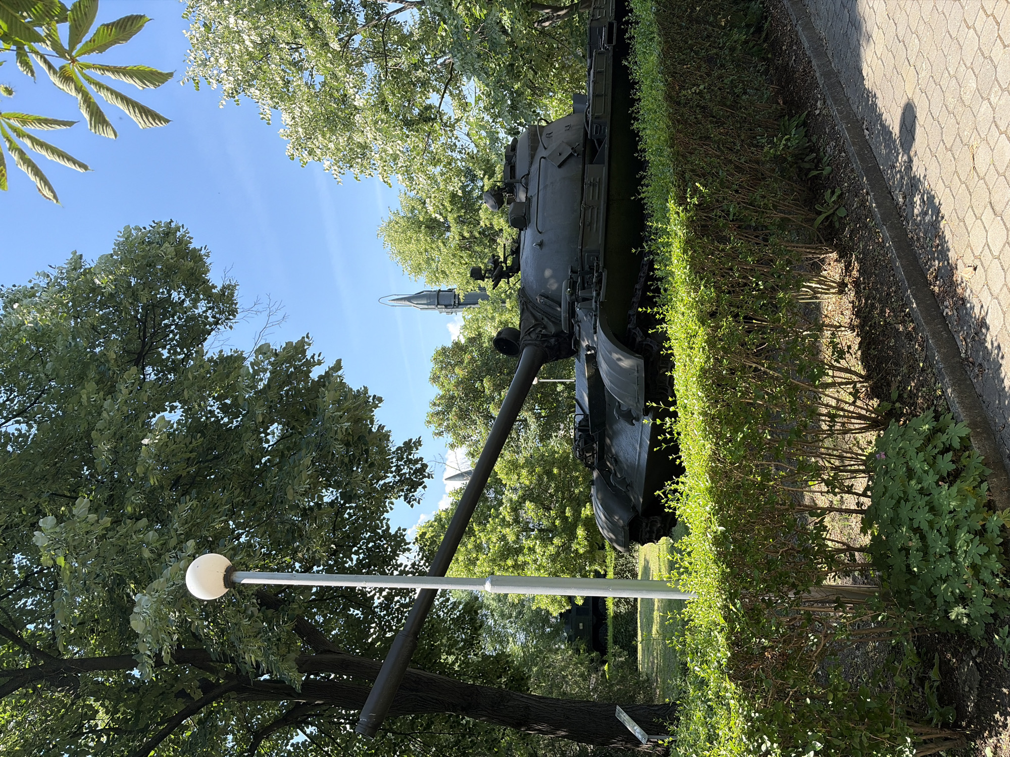 The image depicts a military display in Sofia, Bulgaria. In the foreground, a row of neatly trimmed green bushes lines a paved walkway. Behind the bushes, a dark green tank, likely a museum exhibit, is prominently positioned. Its large cannon extends towards the left of the frame. A tall, slender lamp post with a spherical white light fixture stands to the left of the tank. In the background, lush green trees create a natural backdrop. A tall, slender missile or rocket is visible behind the tank, piercing the bright blue sky. The scene is illuminated by bright sunlight, casting shadows on the walkway and highlighting the vibrant green foliage. There are no people visible in the image.