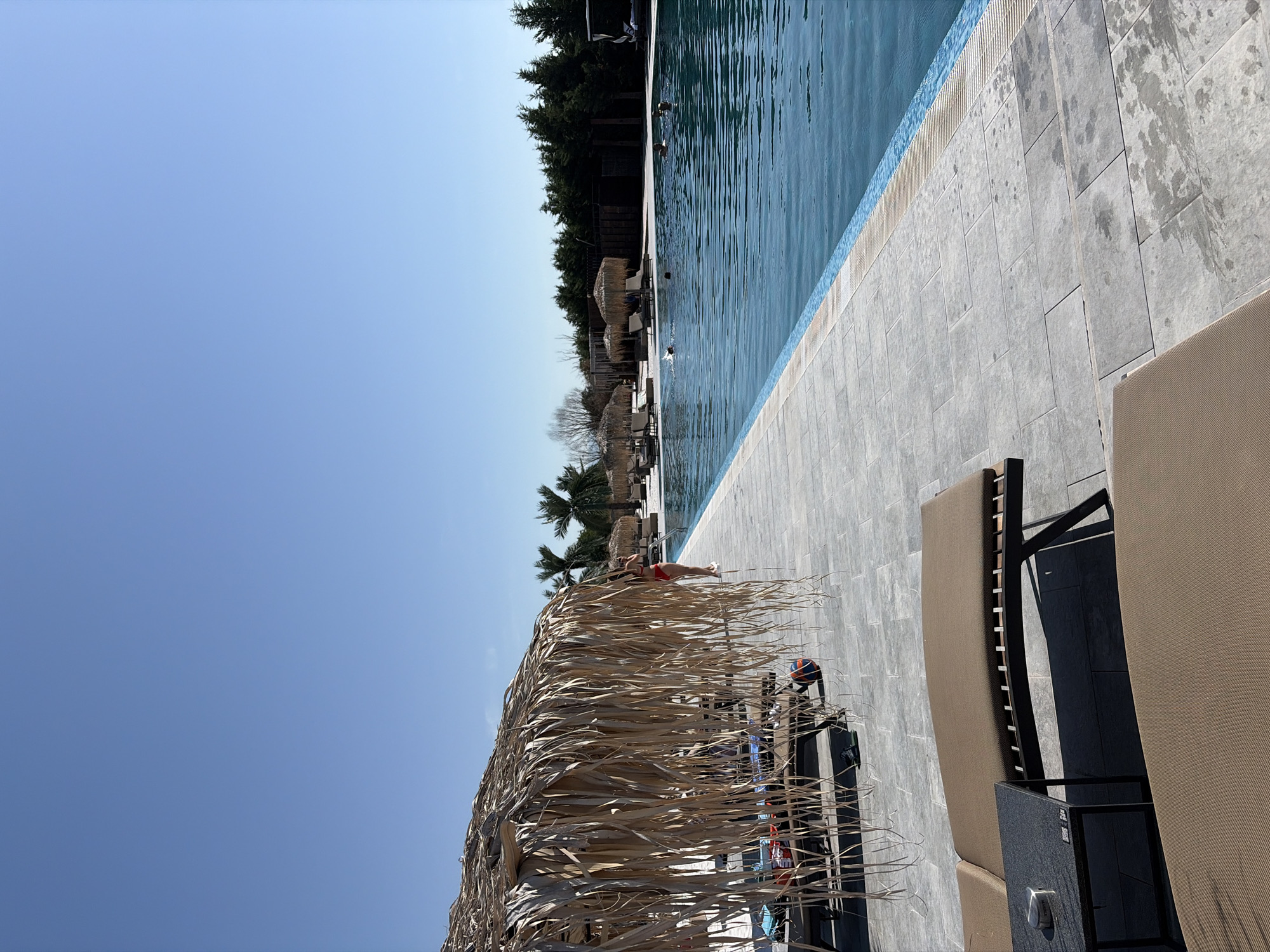 This is a photograph taken at a swimming pool located in Kazichene, Bulgaria. The image captures a sunny day with a clear blue sky. In the foreground, there are several lounge chairs with brown cushions, suggesting a relaxation area. To the left, a thatched umbrella provides shade over some of the lounge chairs, with towels and a small table visible.
The pool itself is large and filled with clear blue water. There are several people in the pool, some swimming and others appearing to be relaxing in the water. Along the edge of the pool, there are more thatched umbrellas and lounge chairs. In the background, a line of dark green trees forms a backdrop, with some additional structures, possibly cabanas or buildings, visible behind them. The paving around the pool is made of light-colored rectangular tiles. The overall scene conveys a sense of leisure and outdoor recreation.