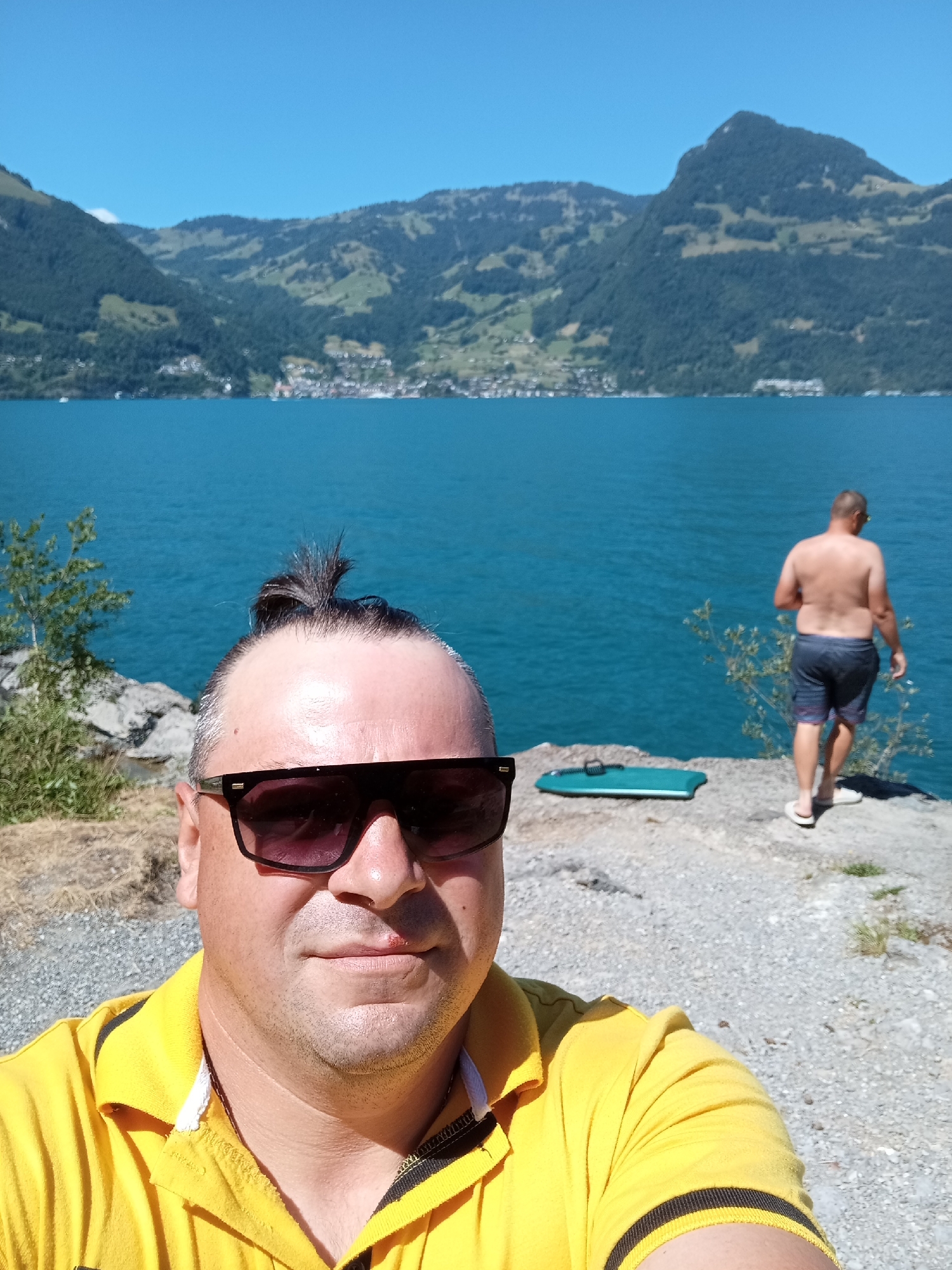 The image shows a man taking a selfie in the foreground, wearing a yellow polo shirt and sunglasses. His hair is pulled up into a small bun. In the background, a body of turquoise water is visible, with mountains and a village on the opposite shore. Another man, shirtless and wearing shorts, stands near the water's edge looking out at the lake. A green flotation device is lying on the rocky ground between the two men. The scene is set in Emmetten, Switzerland, on a sunny day.