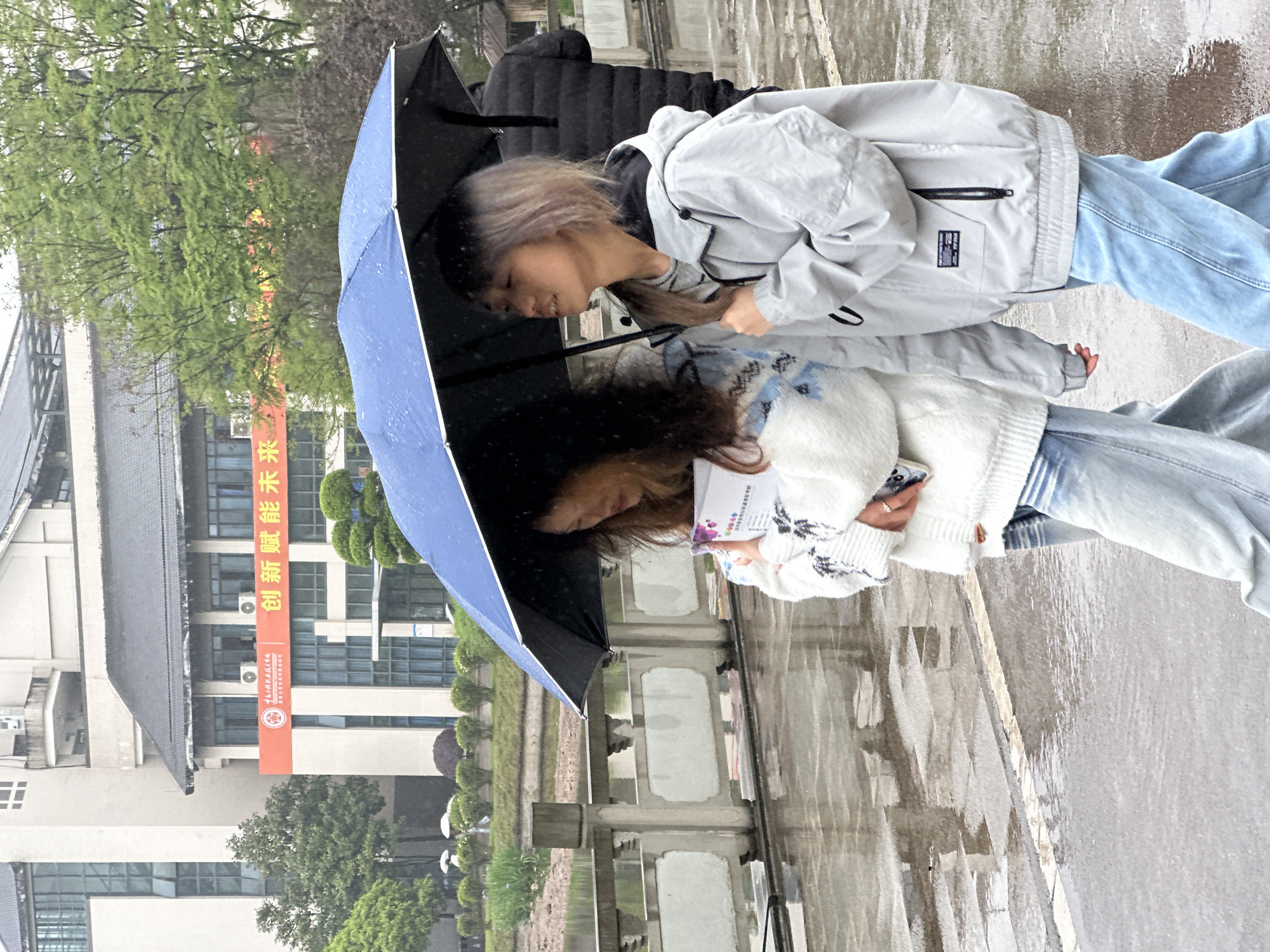 Two women are huddled under a blue and black umbrella on a rainy day. One woman wears a light gray jacket and jeans, the other wears a white sweater and jeans. They are standing next to a stone walkway and railing. In the background is a building with a banner that reads "创新赋能未来" in Chinese, which translates to "Innovation empowers the future." The red logo on the banner indicates it is in the city of Jiangjin, China.