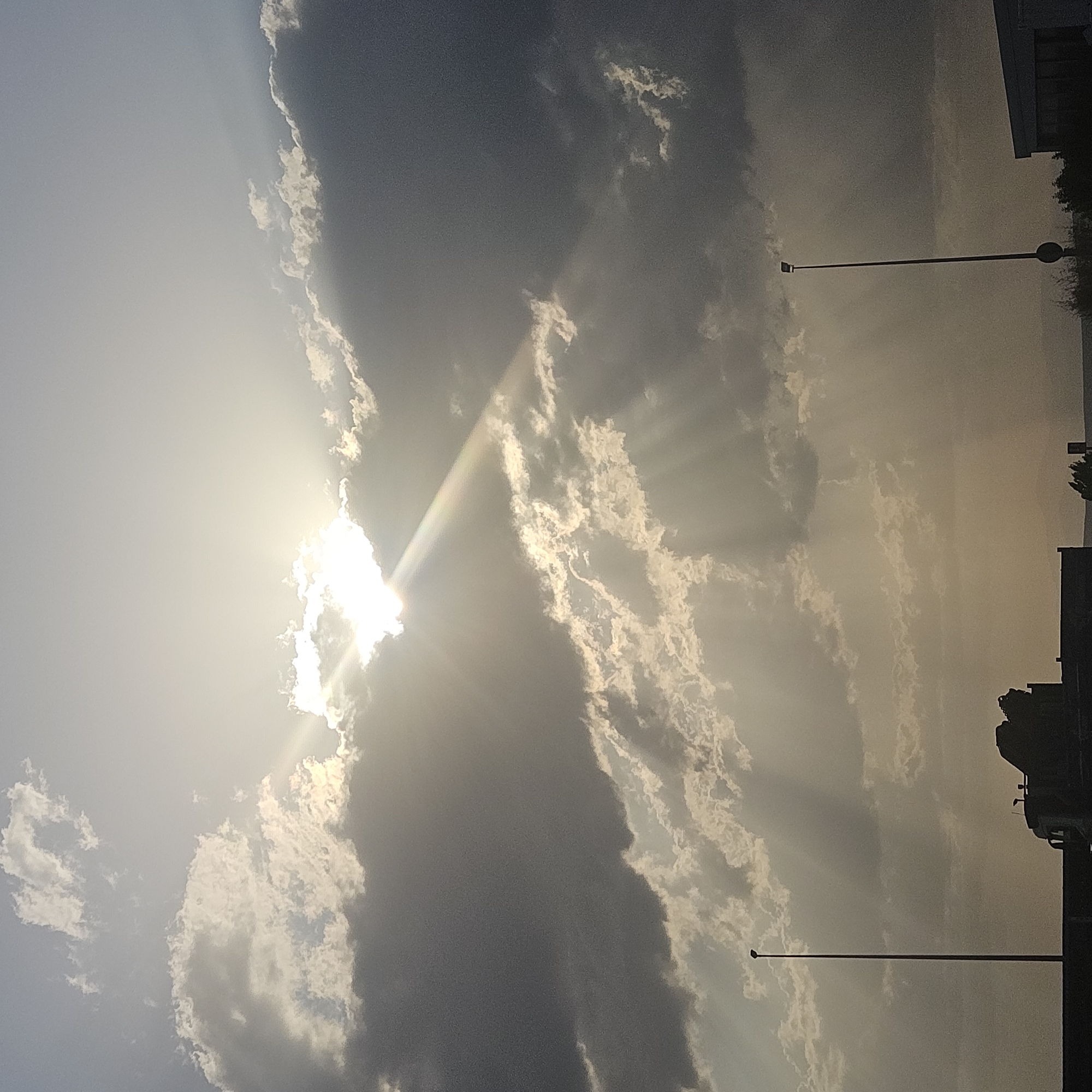 The image shows a cloudy sky with the sun peeking through, casting rays of light. The location is identified as Algora, Spain. In the foreground, there is a silhouette of a vehicle, possibly a truck with a crane, and a tall lamppost. Another lamppost is visible on the right side of the image. The lower part of the image is dark and appears to be the ground or a road. The sky is predominantly filled with grey and white clouds, with areas of brighter light where the sun is visible. The overall scene suggests an outdoor setting, possibly during the daytime, with a hint of industrial or transportation elements due to the vehicle and lampposts.