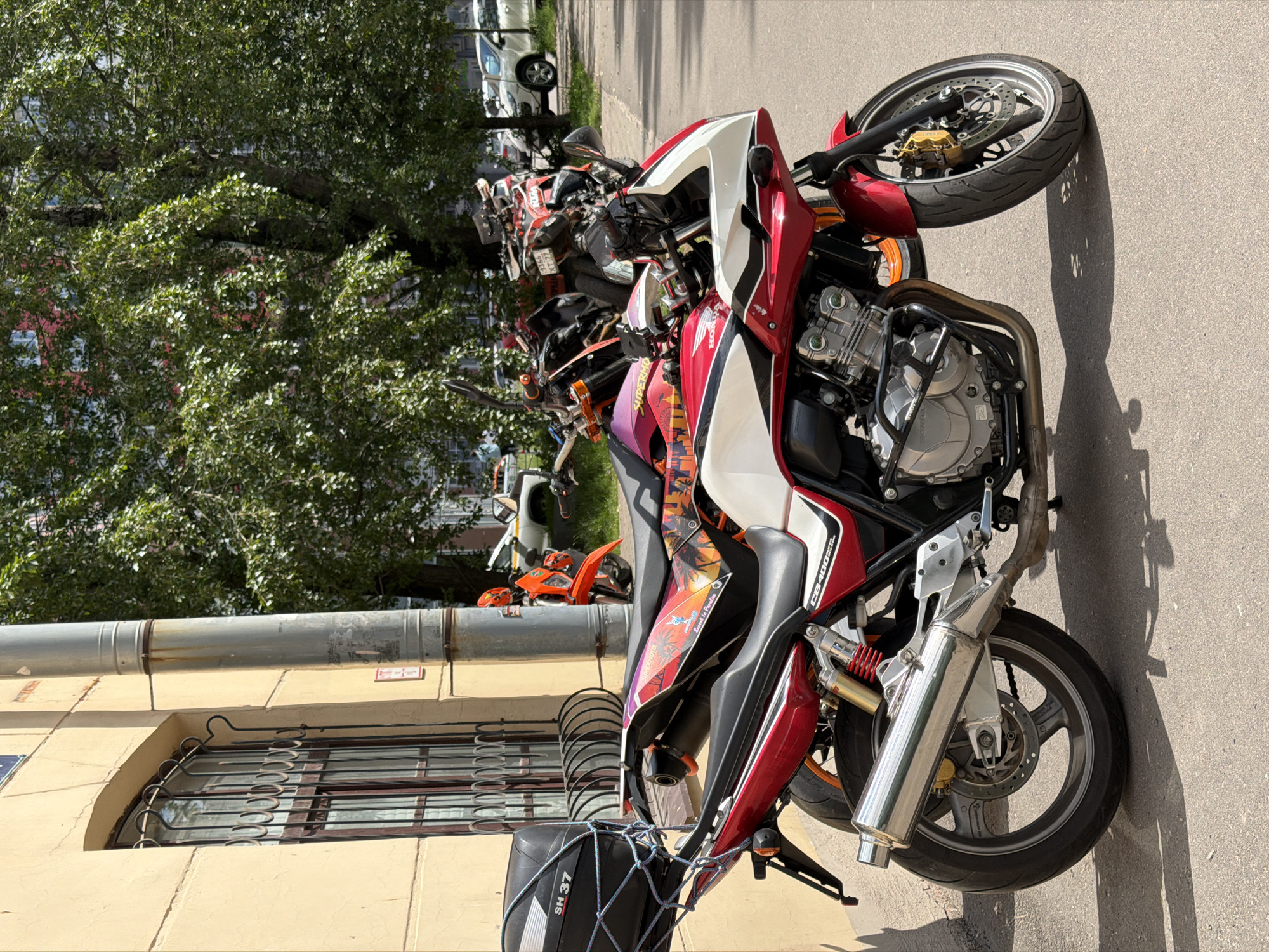 The image captures a street scene in Saint Petersburg, Russia, on a bright, sunny day, featuring several parked motorcycles. The most prominent subject is a red, white, and black Honda CB400 motorcycle, positioned centrally in the foreground.
The Honda CB400 displays "HONDA" branding on its engine cover and "CB400" on its side fairing, which is adorned with various custom stickers, including one that reads "Live life to the fullest." It features a silver exhaust pipe, gold-colored front brake calipers, and a black storage box or bag secured with a net at the rear, bearing the inscription "SH 37."
Behind the foreground motorcycle, several other motorcycles are parked, including an orange one and others with less discernible features. A white car is visible further down the street. One motorcycle's license plate shows partially legible Russian characters, "а166", and another "т 777".
To the left of the motorcycles is a light yellow building with an arched window secured by decorative metal grilles. A large, horizontal corrugated metal pipe is mounted on the building above the window. A lush green tree with dense foliage occupies the upper left portion of the image, casting shadows. The ground is asphalt, marked by the strong, clear shadows of the motorcycles and unseen individuals, indicating bright sunlight, likely midday or early afternoon. The scene suggests an urban setting, possibly a gathering or a common parking area for motorcyclists. A blue street sign is partially visible on the building to the far left.