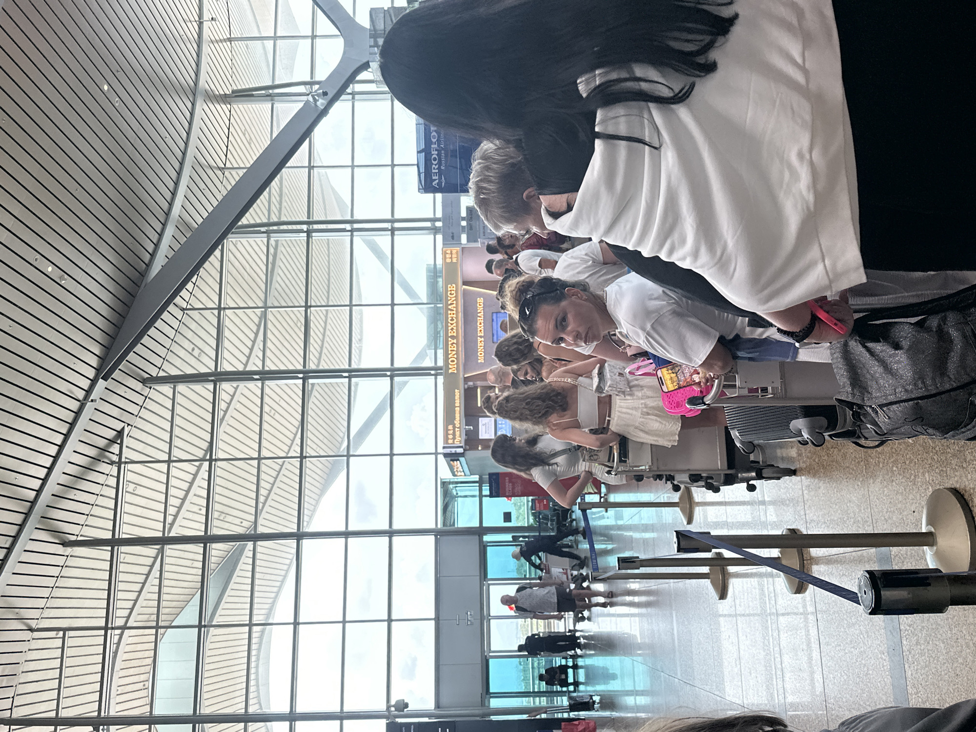 The image shows a crowded scene inside the Cam Ranh International Airport (CXR) located in the city of Bac Cam Ranh Ward, Vietnam. A queue of travelers, mostly wearing casual clothes, are waiting at a service counter, possibly for currency exchange or airline check-in, as indicated by the signs reading "MONEY EXCHANGE" and "AEROFLOT Russian Airline". Some travelers are pulling luggage behind them. There are crowd-control barriers with retractable belts in the foreground. In the background, a large glass window and the airport's ceiling structure with a combination of metal and wooden panels can be observed. Some people are seen walking in the background through the window.