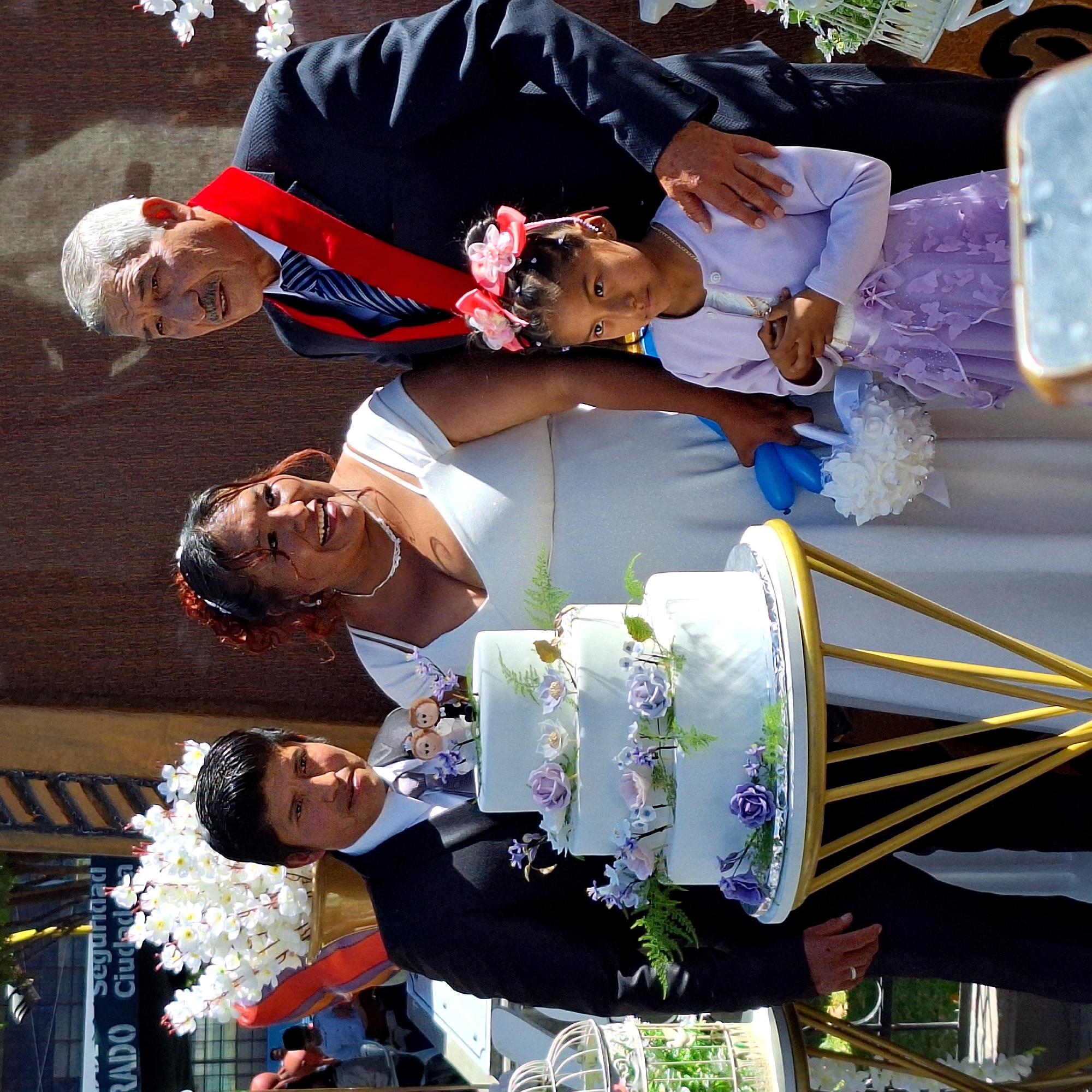 Four people pose with a tiered cake. The backdrop appears to be an urban environment. From left to right: a man with a red sash over his suit, a young girl with a flower hairpiece, a woman in a white dress holding a bouquet, and a man in a suit. A tiered cake is in front of the woman. The image seems to be in Cerro Colorado, Peru.
