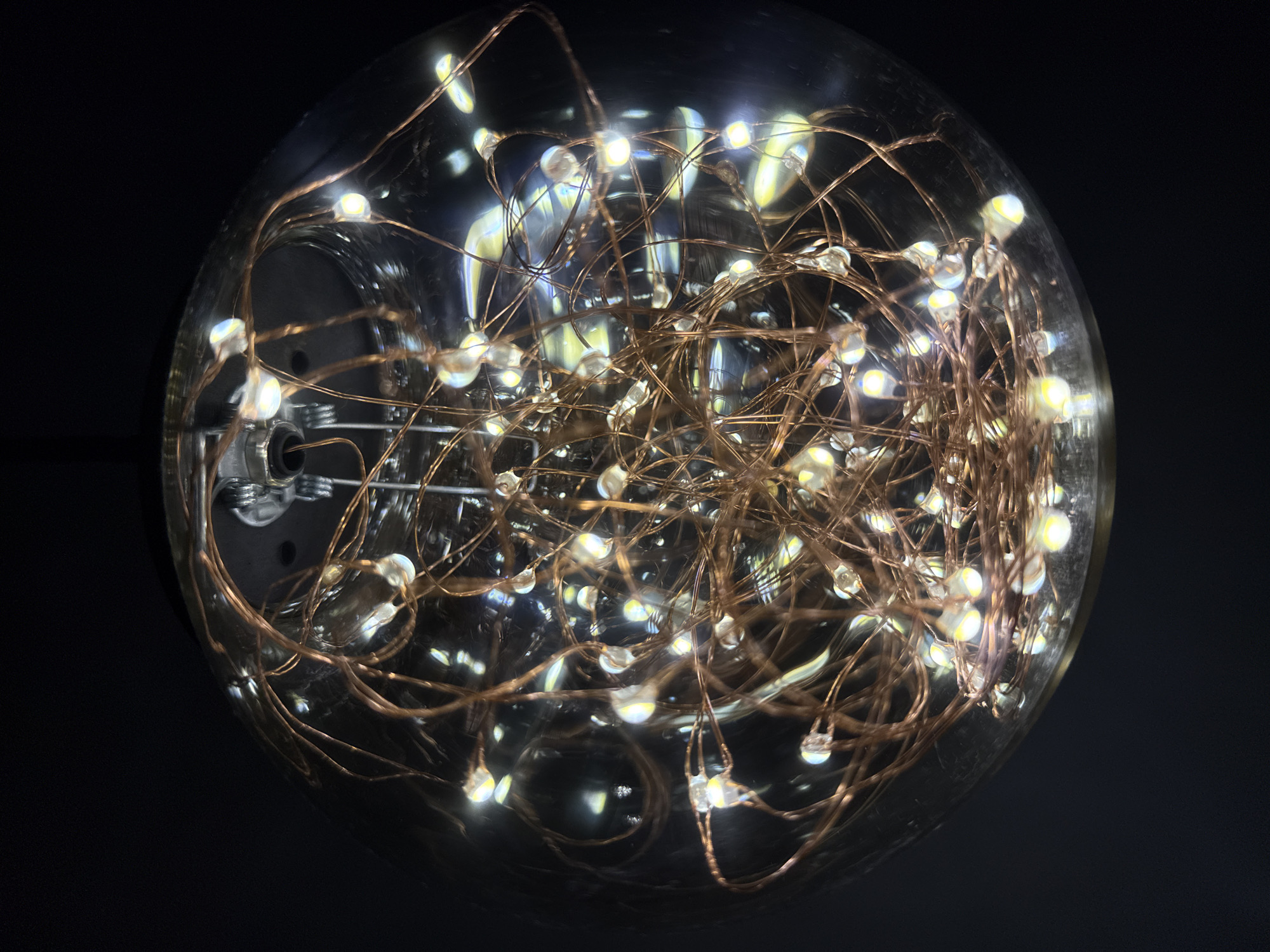 A close-up shot shows a clear glass bulb filled with a dense cluster of small, warm-toned LED lights attached to thin, copper-colored wires. The bulb is against a dark, out-of-focus background, which creates a stark contrast and emphasizes the light. The scene is indoors, with the light from the LEDs illuminating the interior of the bulb and reflecting off the glass. The base of the bulb can be seen at the bottom, with metallic and black components. No people or other subjects are visible. The image captures the soft, diffused light and the intricate arrangement of the wires and LEDs within the bulb. There is no information within the image or supplied that could confirm a location.