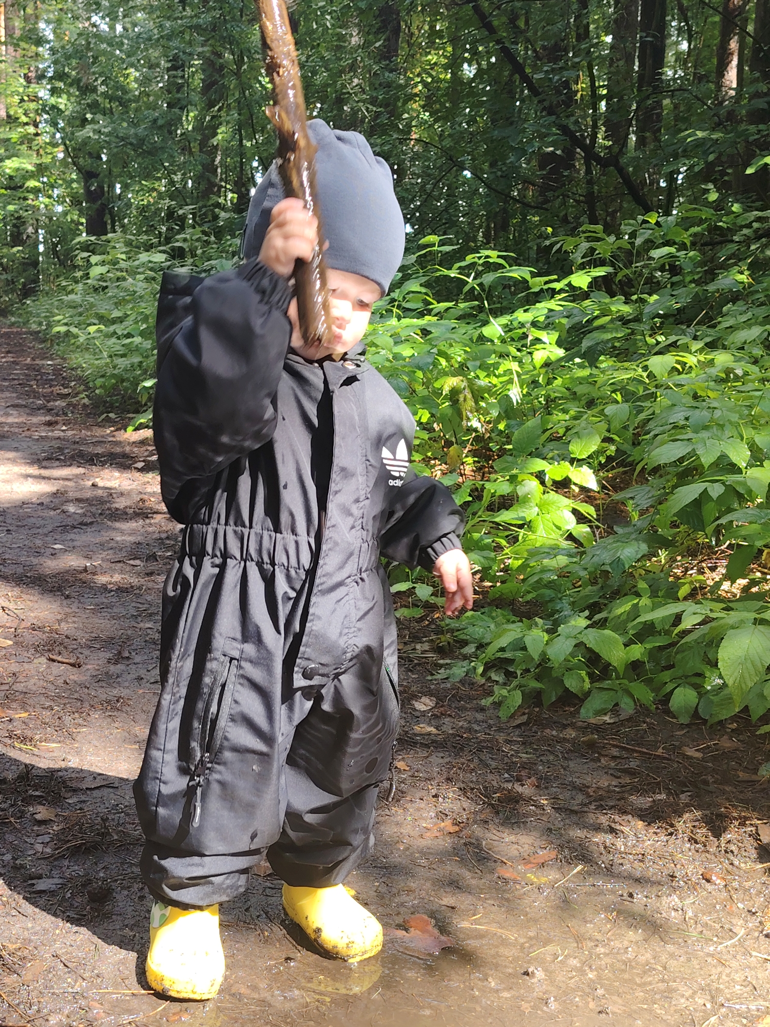 A young child, wearing a black snowsuit and yellow boots, stands in a muddy forest clearing, holding a stick. The child is looking down at the stick. The clearing is surrounded by trees and lush green foliage, suggesting a summer or early autumn setting. The ground is wet and muddy, with a puddle of water in front of the child. The overall scene depicts a child exploring nature, likely in the outskirts of Chaikovsky, Russia, given the context.