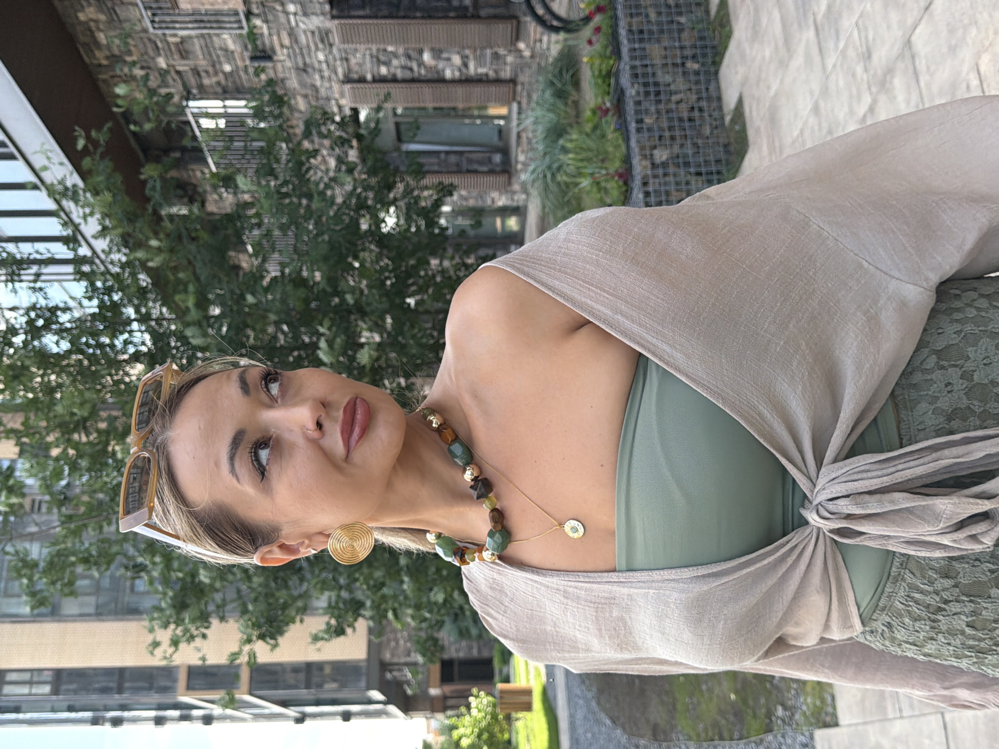 A woman with blonde hair and sunglasses is standing outdoors in front of a building. She is wearing a beige wrap top over a green tube top and a green lace skirt. She has large gold hoop earrings and a chunky multi-colored beaded necklace with a pendant. The background shows a modern brick building and trees. The location is not specified in the image or its metadata.
