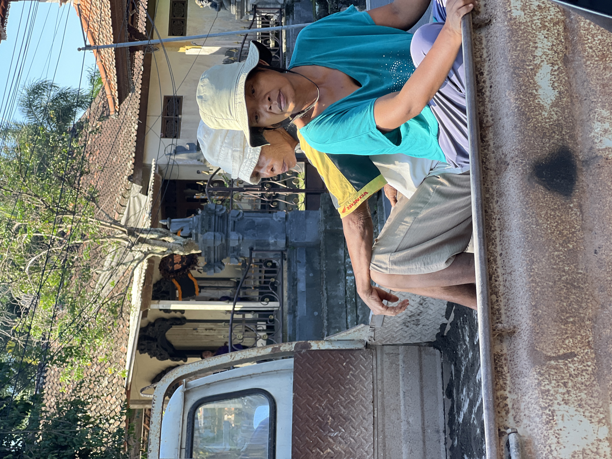 In Sukawati, Indonesia, two individuals, likely of Indonesian descent, are seated in the back of a rusty, weathered white pickup truck. The individual in the foreground, wearing a teal t-shirt and grey pants, is looking forward with a neutral expression. Behind them, partially obscured, is another person wearing a yellow and green shirt and a white hat, also looking forward. The truck bed contains dirt and debris. In the background, a traditional Balinese building with a terracotta tiled roof and ornate architectural details is visible. The scene is illuminated by bright sunlight, casting shadows and highlighting the textures of the truck and the surrounding environment.