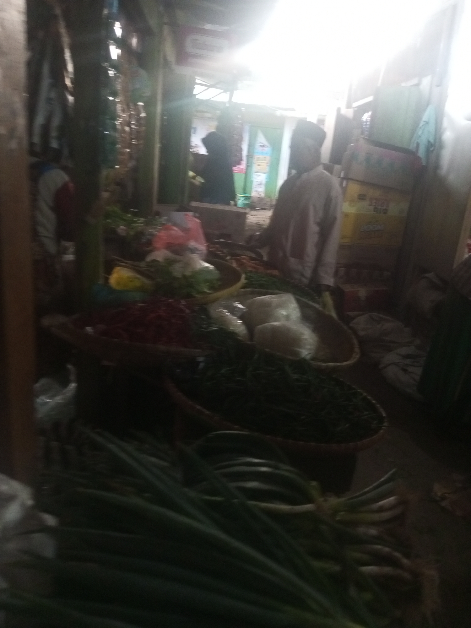 The image depicts a market scene in Kalibeber, Indonesia. In the foreground, a large bundle of green onions is laid out on the ground, with their roots visible. Behind the onions, a woven basket filled with dark green leafy vegetables sits on a dark surface. Further back, another woven basket contains red chili peppers. To the left of the chili peppers, a yellow object, possibly a lemon or lime, is visible. In the middle ground, a man wearing a light-colored shirt and a dark cap stands facing left. He is positioned near a stall where more produce, including what appear to be onions or similar vegetables wrapped in plastic, is displayed in baskets. Behind the man, stacks of cardboard boxes are visible. The background shows a dimly lit interior, with what appears to be a doorway or opening on the left and more stalls and structures on the right. The lighting suggests it might be late in the day or indoors.
