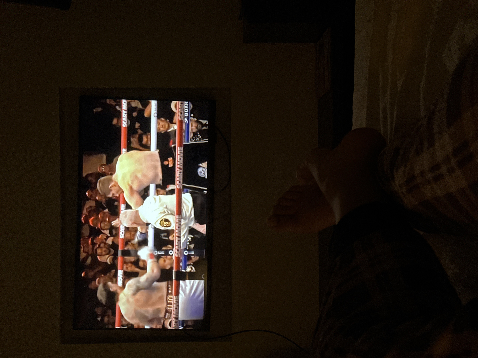 A person is lying in bed, positioned to the right of the frame, with their bare feet visible. Their legs are covered by dark pants and a flannel-patterned shirt. To the left, a television screen displays a boxing match with a crowd in the background. The boxing ring is indicated by ropes labeled "Scary Movie Boxing." The setting appears to be indoors, possibly a bedroom. There is no direct evidence within the image to confirm or refute a location in Genk, Belgium.