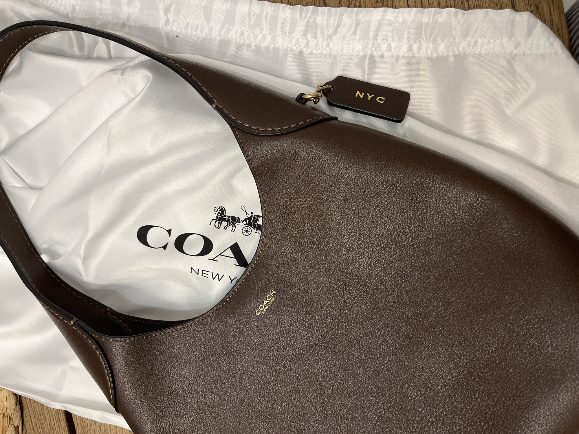 A brown leather Coach shoulder bag is displayed on a white fabric dust bag, which is laid on a wooden surface. The dust bag has the Coach logo printed on it in black. A dark brown leather luggage tag with "NYC" embossed in gold lettering is attached to the bag's strap. The bag has a textured leather finish and visible stitching. The scene is lit by artificial light. There are no people or animals present in the image, and the setting appears to be indoors. The location in Limassol, Cyprus, is not discernible from the image itself.