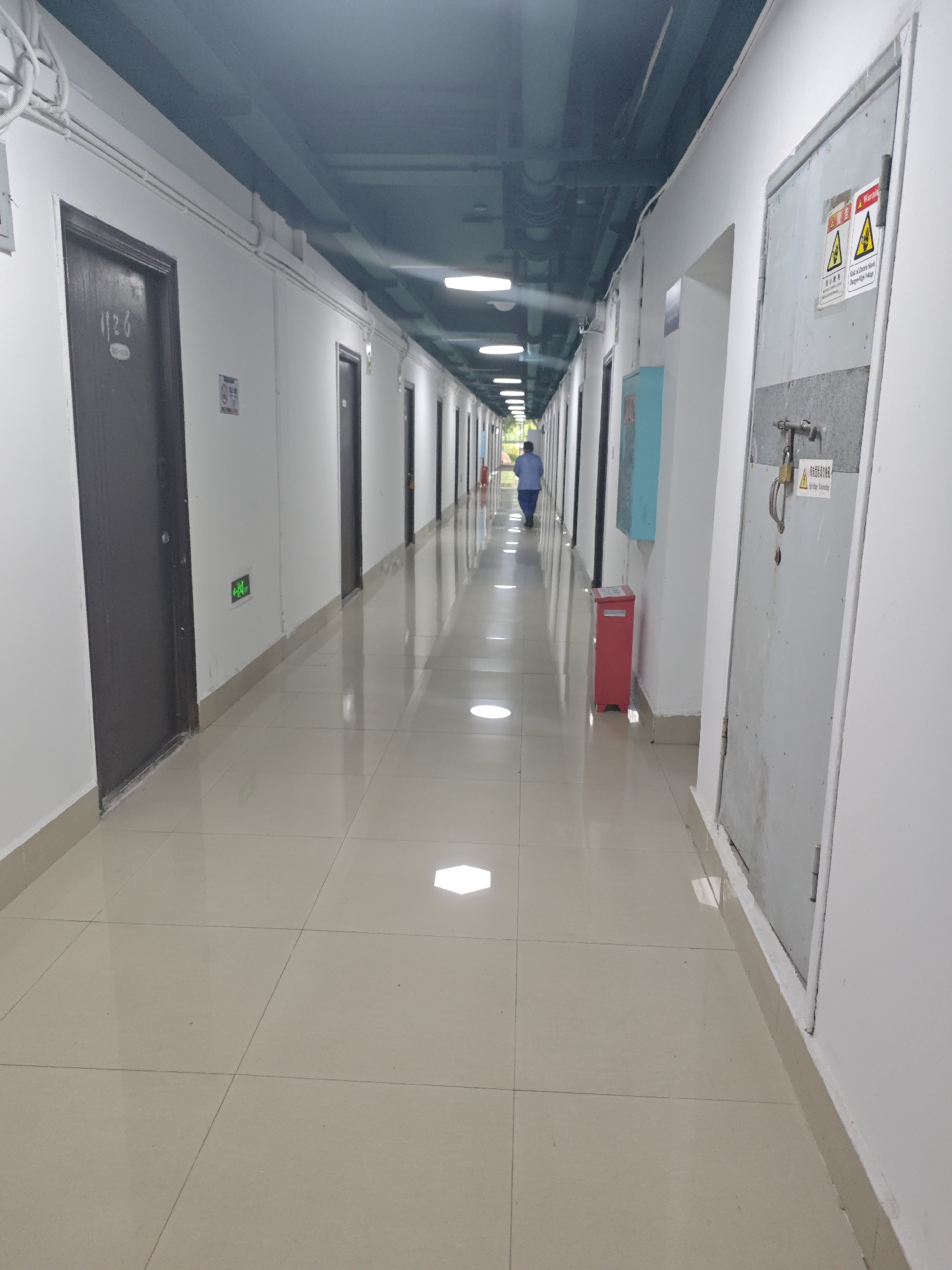 The image shows a long, brightly lit hallway with doors on either side. The hallway appears to be in a building located in the Chongqing High-tech Zone, China. The walls are painted white, and the floor is tiled with a light-colored, reflective material. Overhead, there are fluorescent lights and exposed pipes and conduits.
On the left side of the hallway, there are several dark-colored doors with room numbers visible on some of them. One door has the number "1126" on it. There is also a green illuminated exit sign on the wall between two doors. Further down the hallway, a person in a blue shirt is walking away from the camera.
On the right side of the hallway, there is a large, gray metal door with a padlock and warning signs. Below this door, a bright red trash receptacle is visible. Further down the right wall, a light blue cabinet is mounted on the wall. The overall impression is of a functional, indoor environment, likely a corridor in an office building or dormitory.