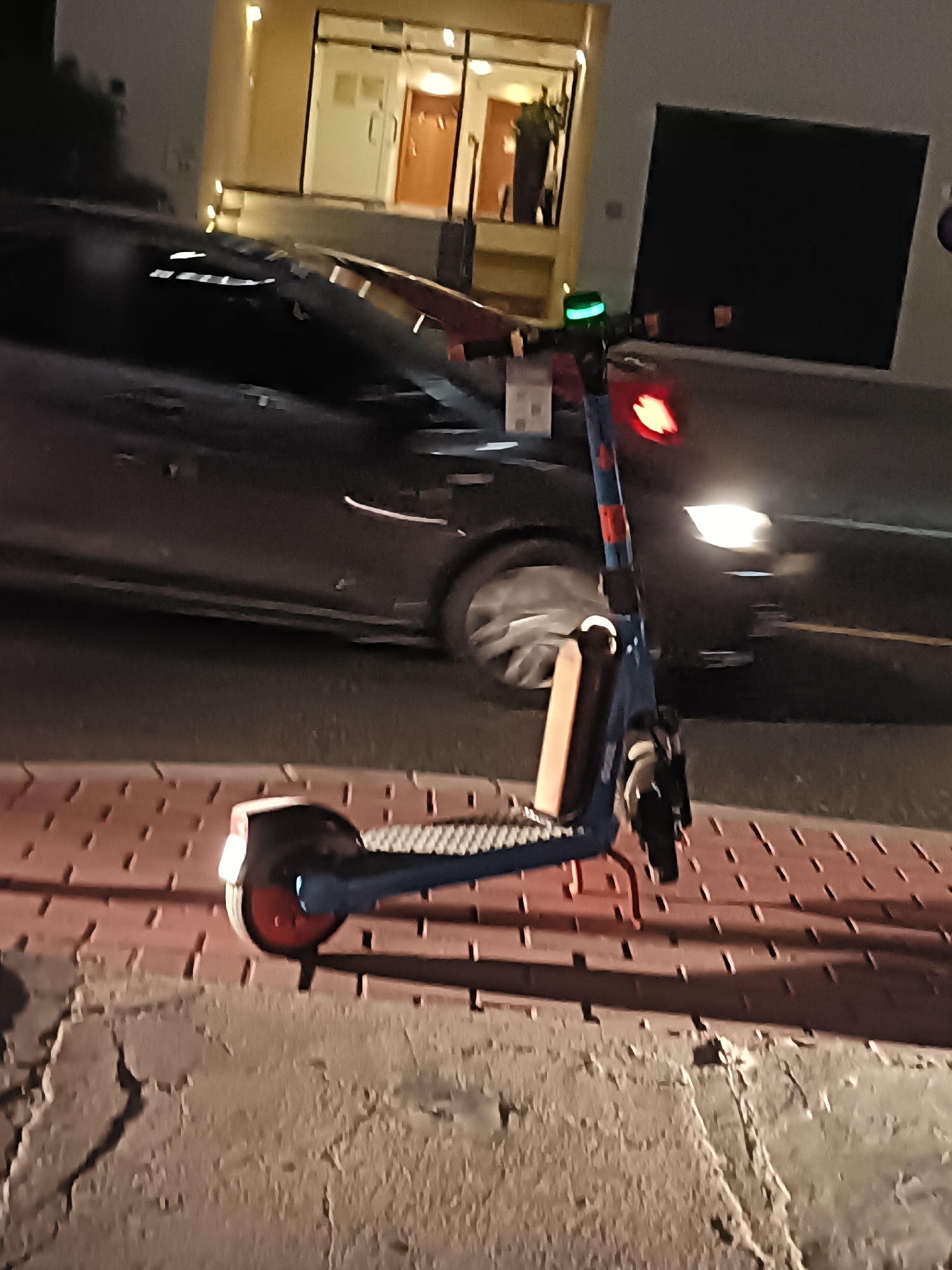 A blue electric scooter with an orange wheel and a red brake light is parked on a red tiled sidewalk at night. The scooter has a green light on its handlebar. Behind the scooter, a dark-colored car is visible, with its headlights on and reflecting on the wet asphalt. The background shows a building with glass doors and windows, illuminated by warm light. The scene appears to be a street in Dubai, United Arab Emirates, given the architectural style of the building and the presence of electric scooters, which are common in the city. The pavement is made of interlocking red tiles, and there is a cracked concrete wall in the foreground.
