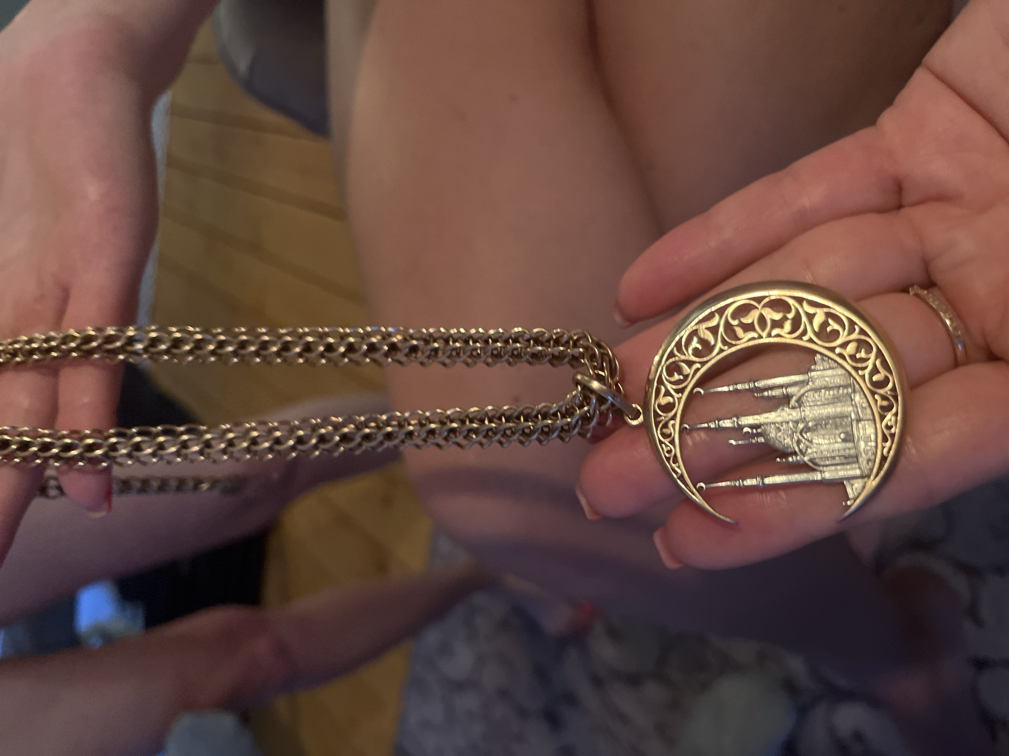 A person is holding a gold-colored necklace with a crescent moon pendant in their hand. The pendant features a detailed etching of a structure resembling a mosque or palace within the crescent shape. The necklace chain is thick and made of interlocking links. A ring is visible on the person's finger. The background is blurry, showing a combination of skin tones and clothing. There is no indication of the location being Новая Чернушка, Russia, in the image.