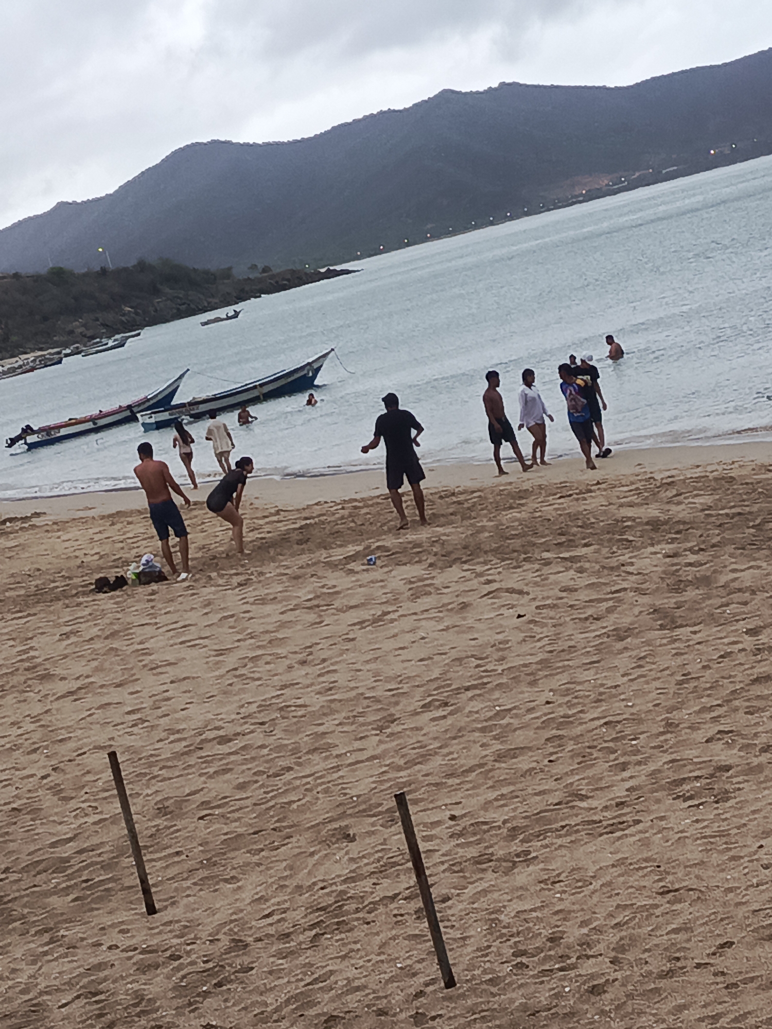 The image depicts a beach scene in Juan Griego, Venezuela, under an overcast sky. In the foreground, a sandy beach is visible with two dark poles embedded in the sand. Several individuals are present on the beach and in the shallow water. On the sand, a man in dark shorts and a shirtless torso stands, facing away from the camera, with items scattered at his feet. A woman in dark athletic wear is crouched down. Further back, closer to the water's edge, a group of people are interacting. Some are in the water, while others are on the wet sand.
In the middle ground, two motorboats are partially submerged in the water, tied to the shore. The water is a pale blue-green. In the background, a body of water stretches towards a hilly, mountainous coastline. The slopes of the hills are covered in vegetation, and a few lights are visible along the distant shoreline, suggesting human settlement. The overall atmosphere is muted due to the cloudy weather.