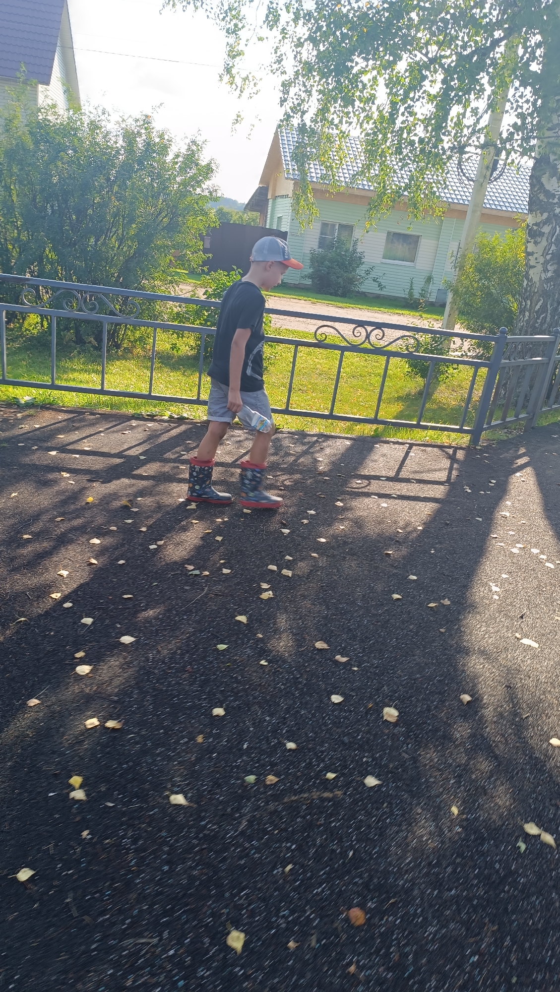 A young boy wearing a black t-shirt, grey shorts, a grey baseball cap with an orange brim, and colorful rubber boots walks on a paved surface outdoors. He is holding a white bottle in his right hand. The ground is dark asphalt with scattered fallen leaves. A black metal fence with decorative scrollwork runs along the left side of the image, separating the paved area from a grassy strip. Behind the fence, a light green building with a dark roof is visible, surrounded by trees and vegetation. The sun is casting shadows on the ground, indicating it is either morning or late afternoon. The scene appears to be a residential area, possibly a yard or a pathway near houses.