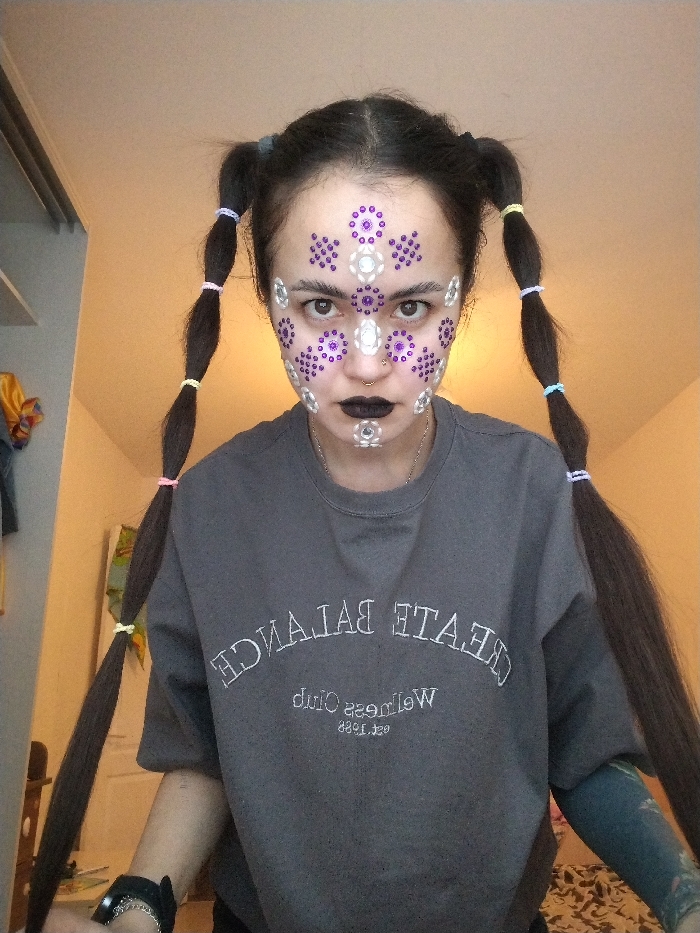 A young individual is depicted from the waist up, centered in an indoor setting in Noviy, Russia. The person, appearing female, has dark hair styled into two long pigtails segmented with multi-colored elastic bands (pink, yellow, light blue, purple). Her face is adorned with numerous adhesive rhinestones; silver gems highlight the brow, nose bridge, and chin, while purple gems are clustered on the forehead and cheeks. She wears a silver septum ring and dark lipstick. Her attire consists of a dark gray short-sleeved sweatshirt featuring inverted white embroidered text that reads "CREATE BALANCE Wellness Club est. 1988". A dark wristband with a chain is visible on the left wrist, and a light-colored tattoo is partially visible on the right forearm. The background consists of a plain, light-colored wall with a partial view of a white shelving unit or closet on the left. The subject is directly facing the camera with a neutral expression.
