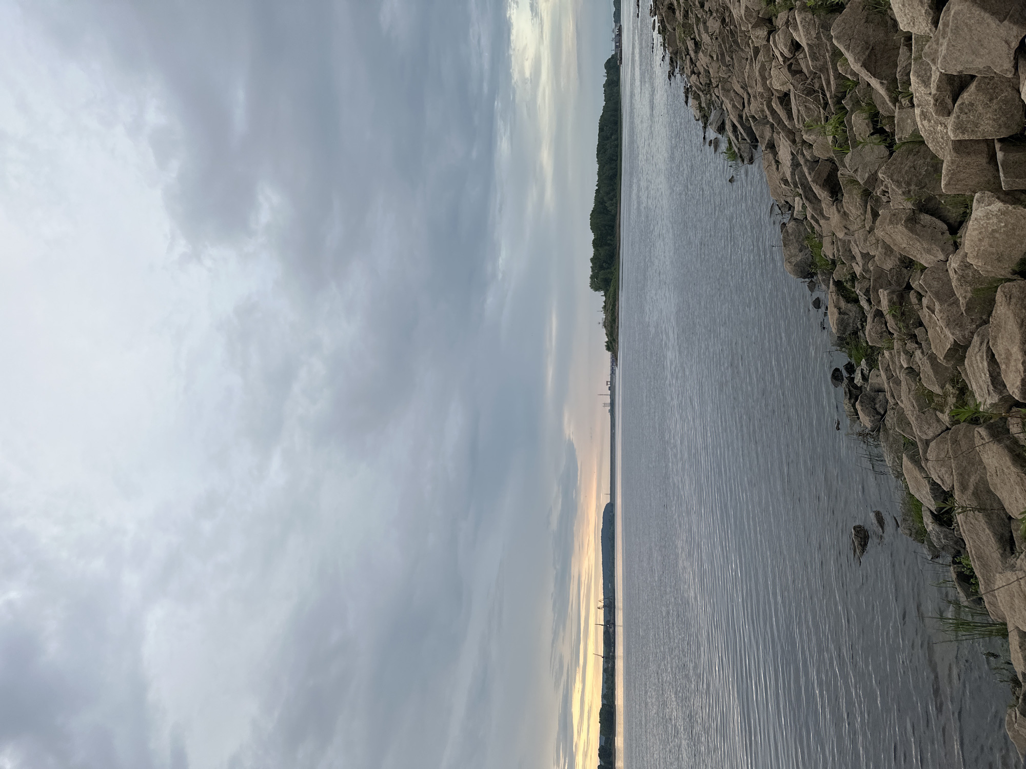 This media file captures a view of a wide river from a rocky embankment. The scene is dominated by a large body of water, with ripples suggesting a gentle breeze. On the right side of the image, the riverbank is densely packed with large, gray rocks of varying sizes, interspersed with patches of green grass and weeds. The water extends to the horizon, where a distant tree line forms a dark green border against the sky.
The sky is predominantly filled with clouds, which are heavy and a mix of gray and white, indicating overcast or cloudy weather. Towards the horizon, there's a softer, brighter band of light, suggesting either early morning or late evening. Faint structures, possibly cranes and buildings, can be seen along the distant shoreline, hinting at an industrial or port area. The overall mood is serene and natural, with the rock formations providing a rugged texture against the smooth expanse of the water and sky. There is no visible text in the image. The lighting suggests it is either dawn or dusk.
