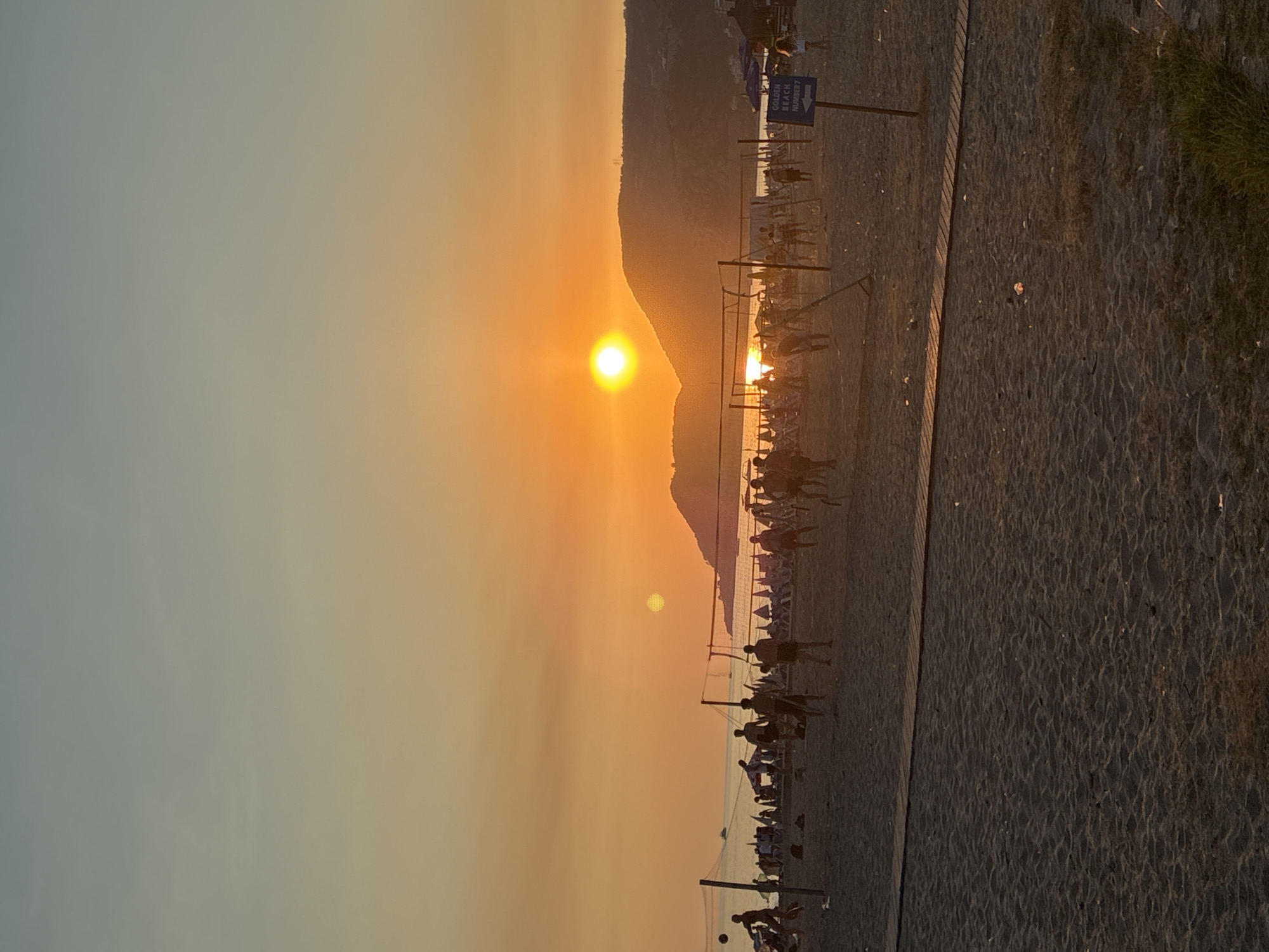 The image shows a beach scene at sunset, likely in Alanya, Turkey, given the presence of volleyball games. The sun is setting over a body of water, casting a warm orange glow across the sky and beach. A group of people are playing volleyball on the sand, silhouetted against the bright sky. In the background, there are beach umbrellas, lounge chairs, and a few boats. A wooden walkway is laid out on the sand, leading towards the volleyball court. The overall atmosphere is one of leisure and recreation.